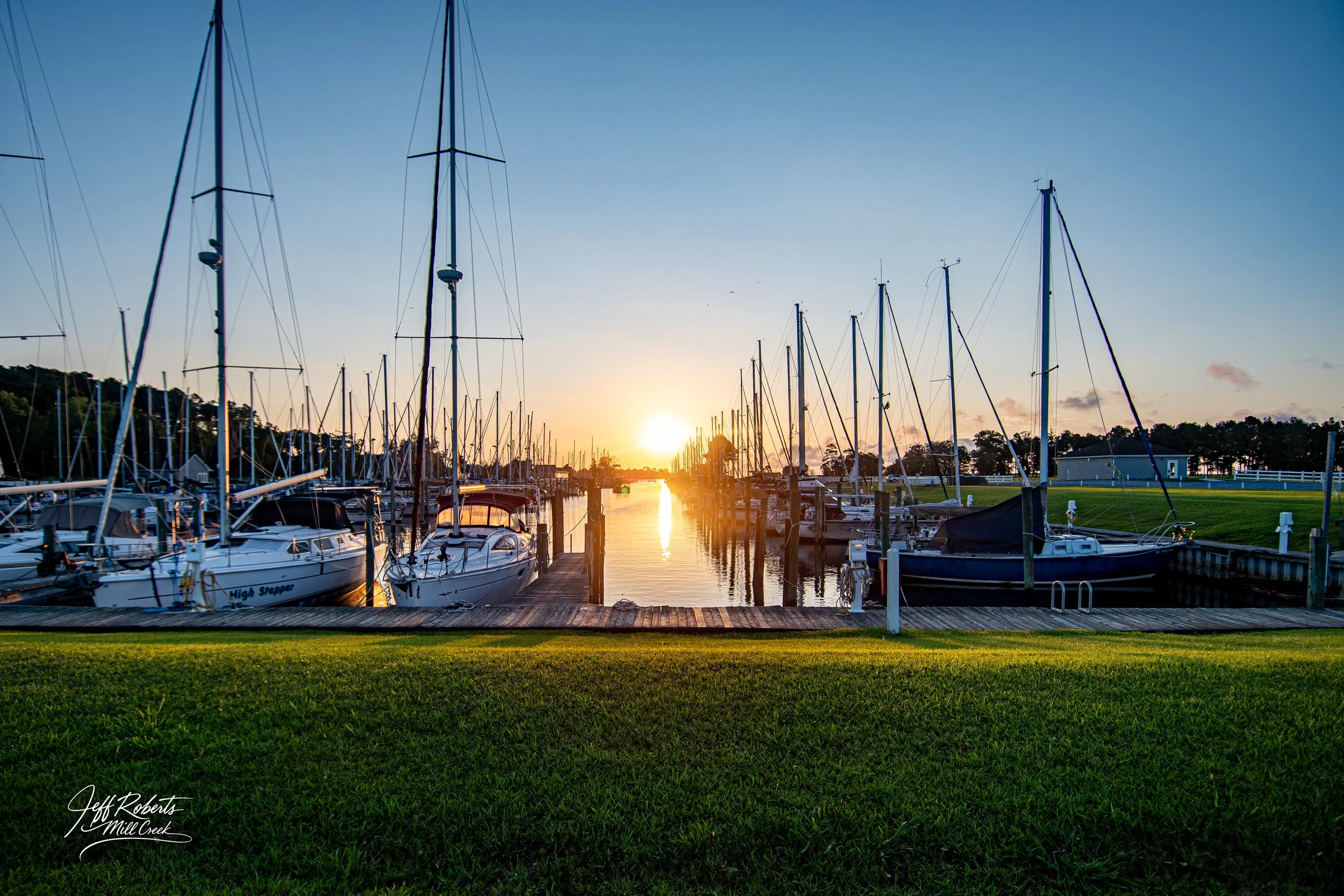 Sailboats docked at a marina during sunset with a grassy foreground and a calm waterway.