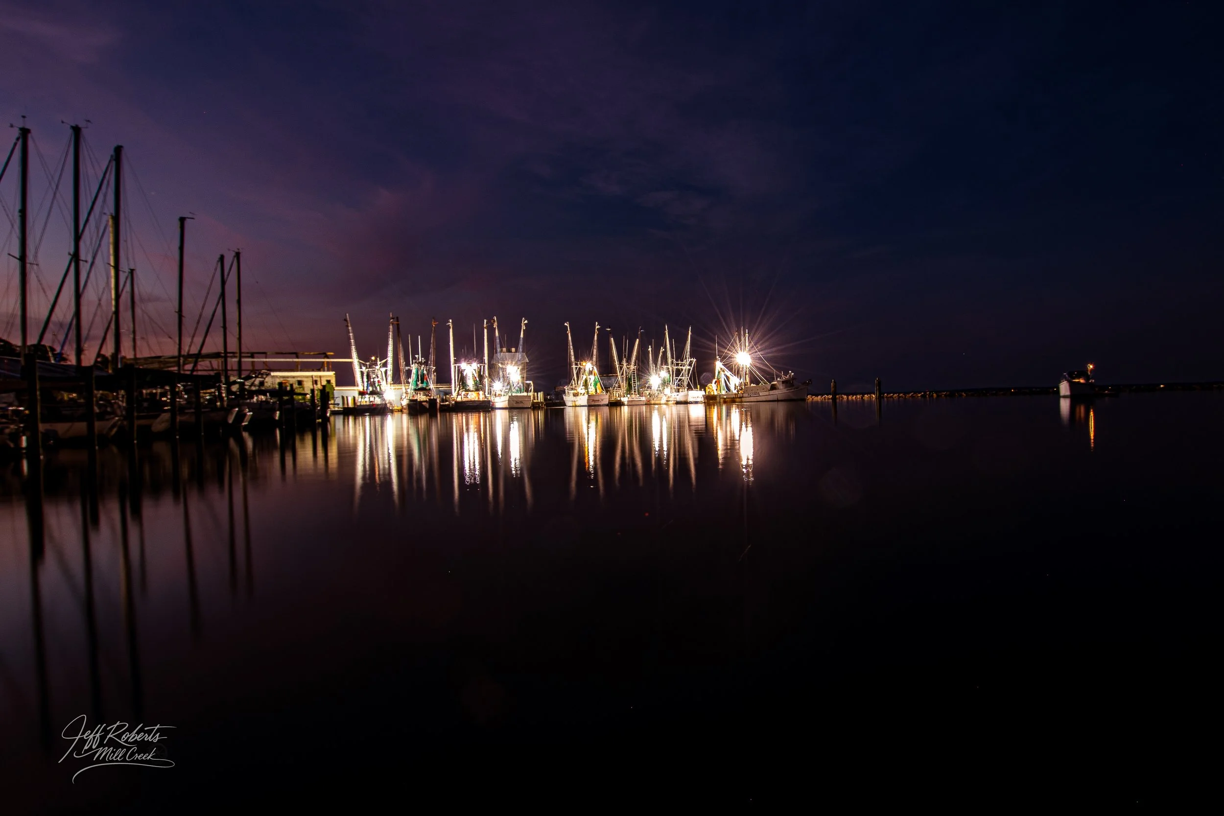 Nighttime view of a marina with sailboats and yachts, reflections on calm water, dark sky with some clouds, bright lights on boats creating starburst effects, signature in bottom left corner.