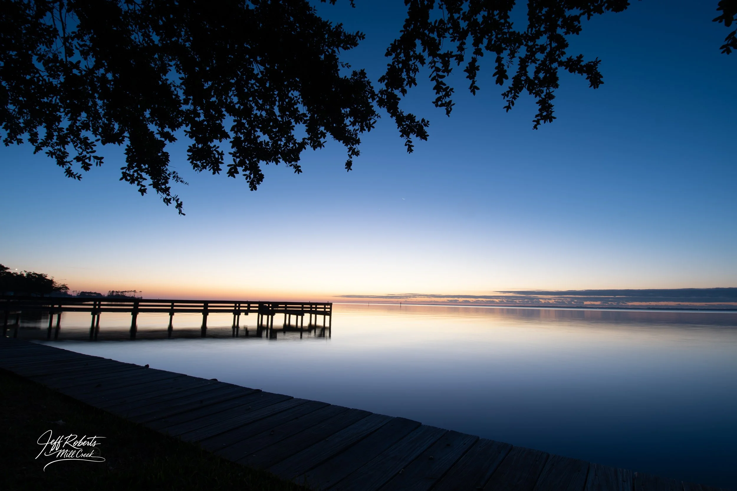 A peaceful lake at sunset with a wooden dock extending into the water. Silhouettes of tree branches frame the scene at the top, and the sky transitions from orange near the horizon to deep blue overhead. Reflective water mirrors the colors of the sky