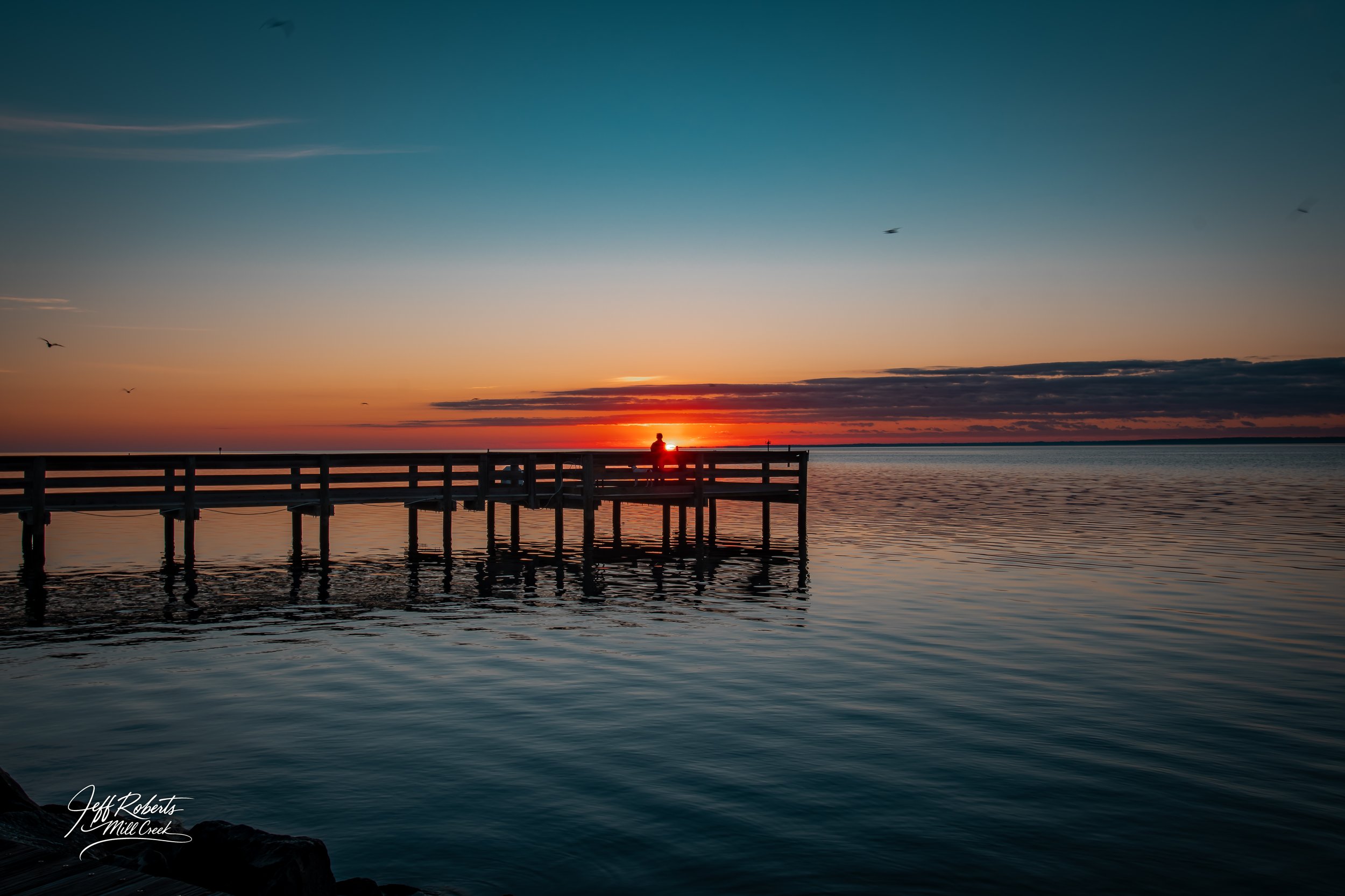 Sunset over a calm lake with a wooden pier and a person sitting at the end of the pier, clouds and birds in the sky.