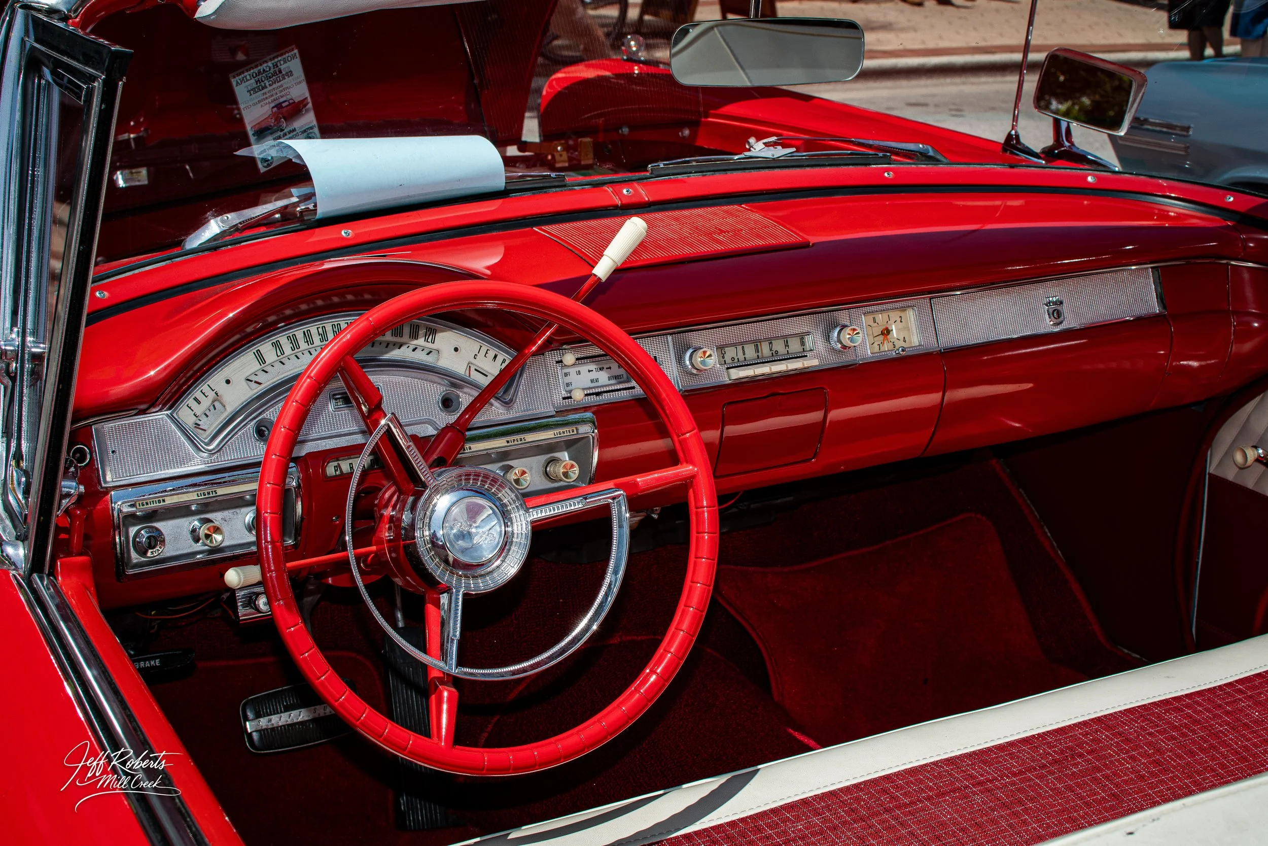 Interior of a vintage red convertible car, showing a dashboard with a large speedometer, radio, and controls, and a red steering wheel with a chrome center.