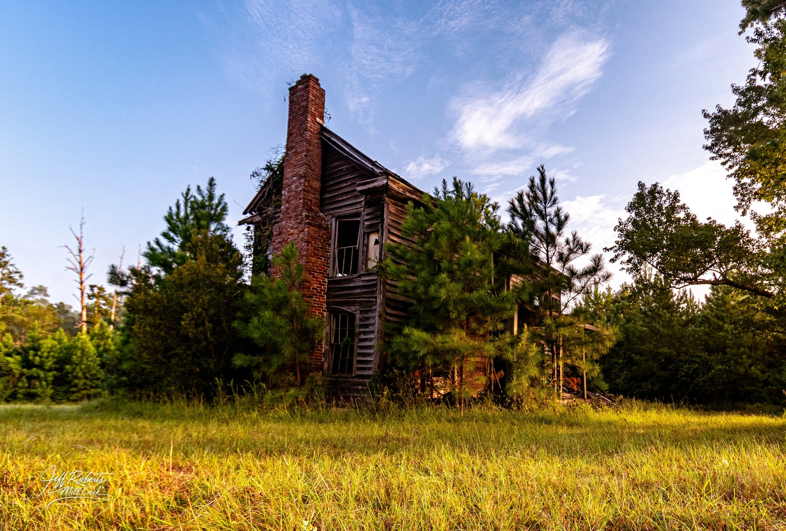 An old, abandoned two-story wooden house with a brick chimney, overgrown with trees and grass, under a partly cloudy sky at sunset.