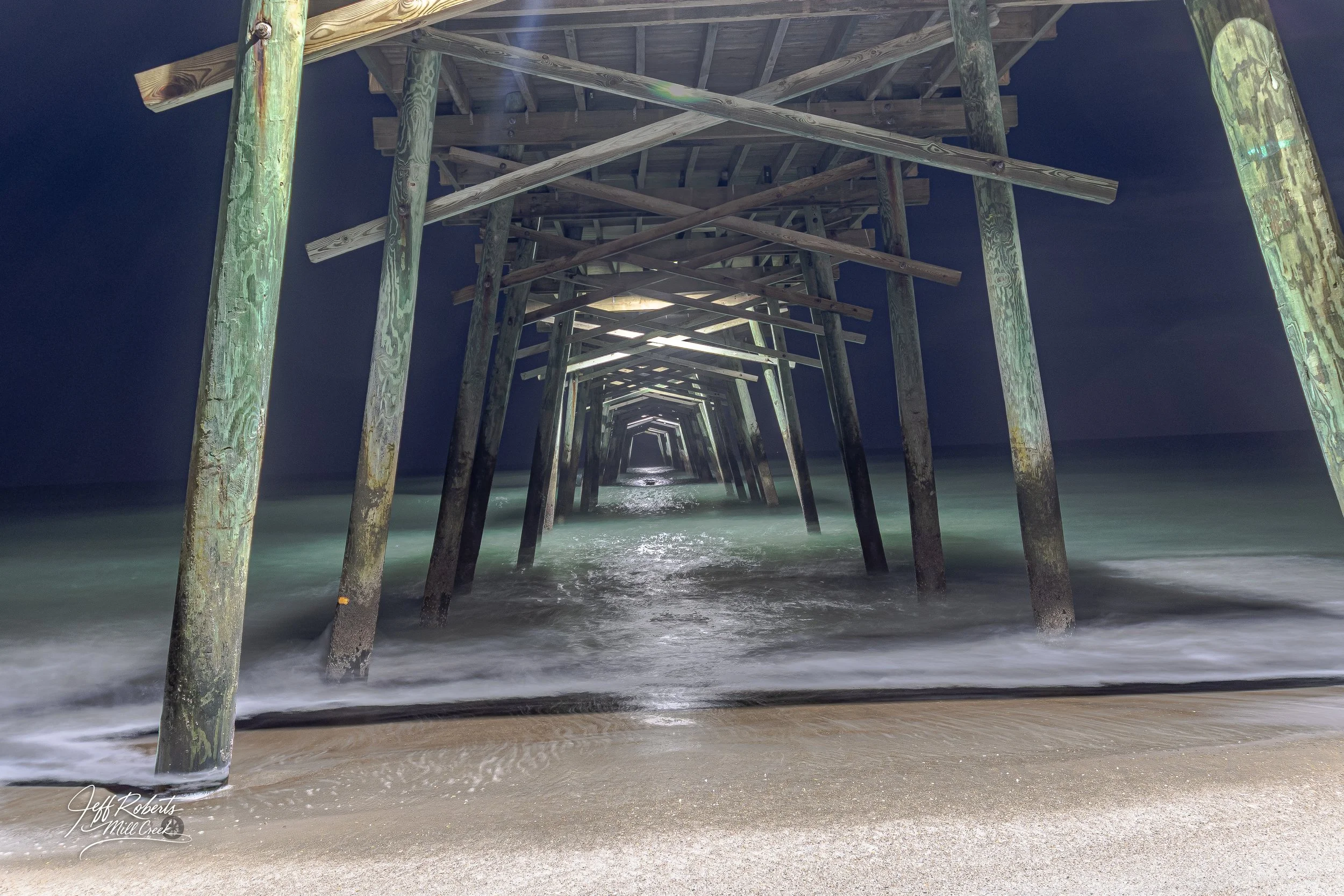 View from the beach of a wooden pier extending into the ocean at night, with underwater support beams visible and gentle waves at the shore.