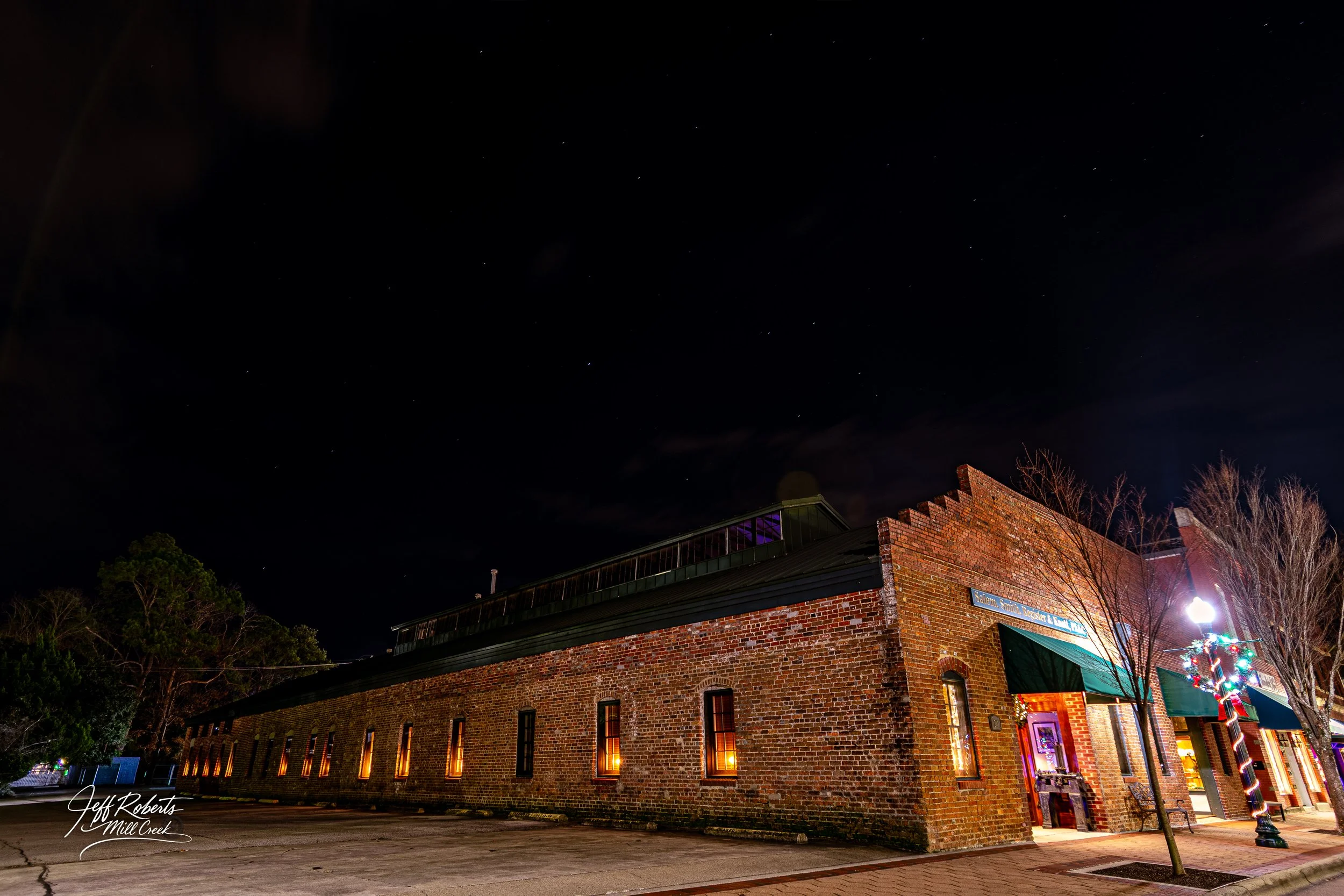 Night view of a brick building with green awnings and lit windows, decorated with Christmas lights on a lamppost in a small town.