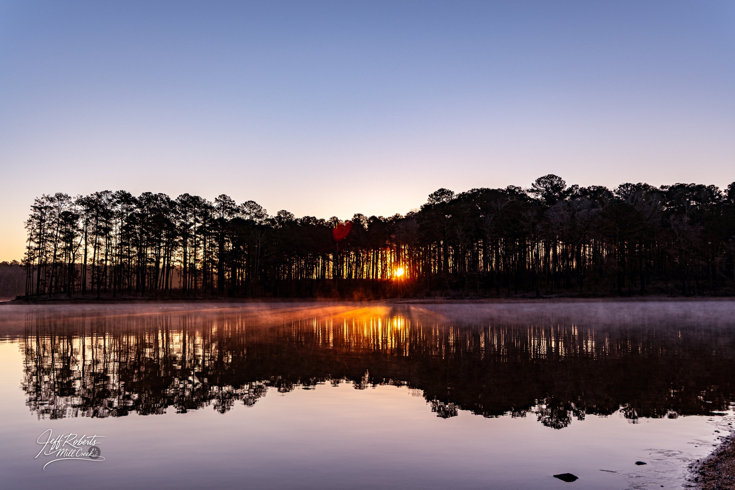 Sunset over a calm lake with a tree-lined shoreline, reflections in the water, and mist rising from the surface.