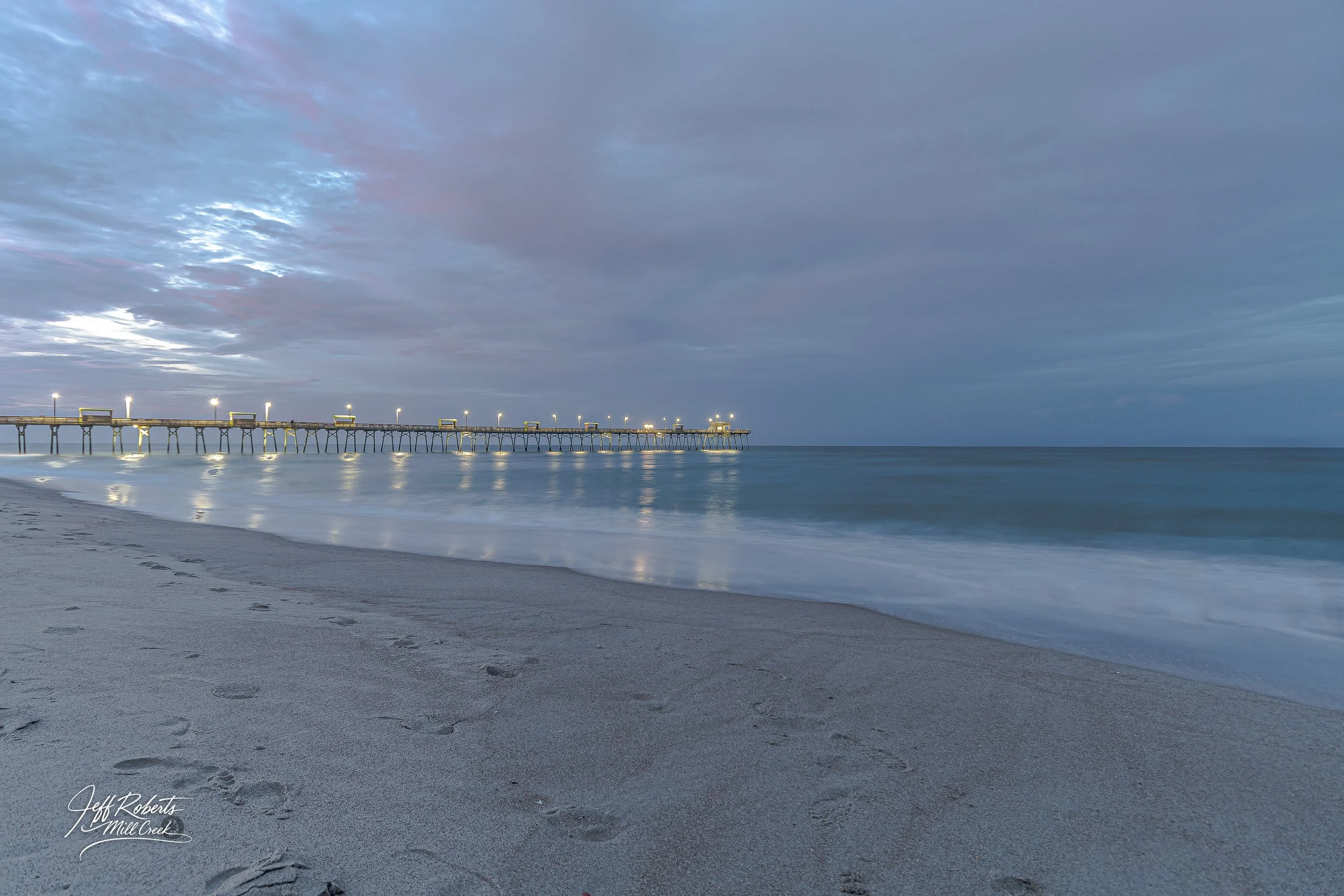 Long pier extending into the ocean with lights on its posts, set against a cloudy sky during dawn or dusk, with calm waves on a sandy beach in the foreground.