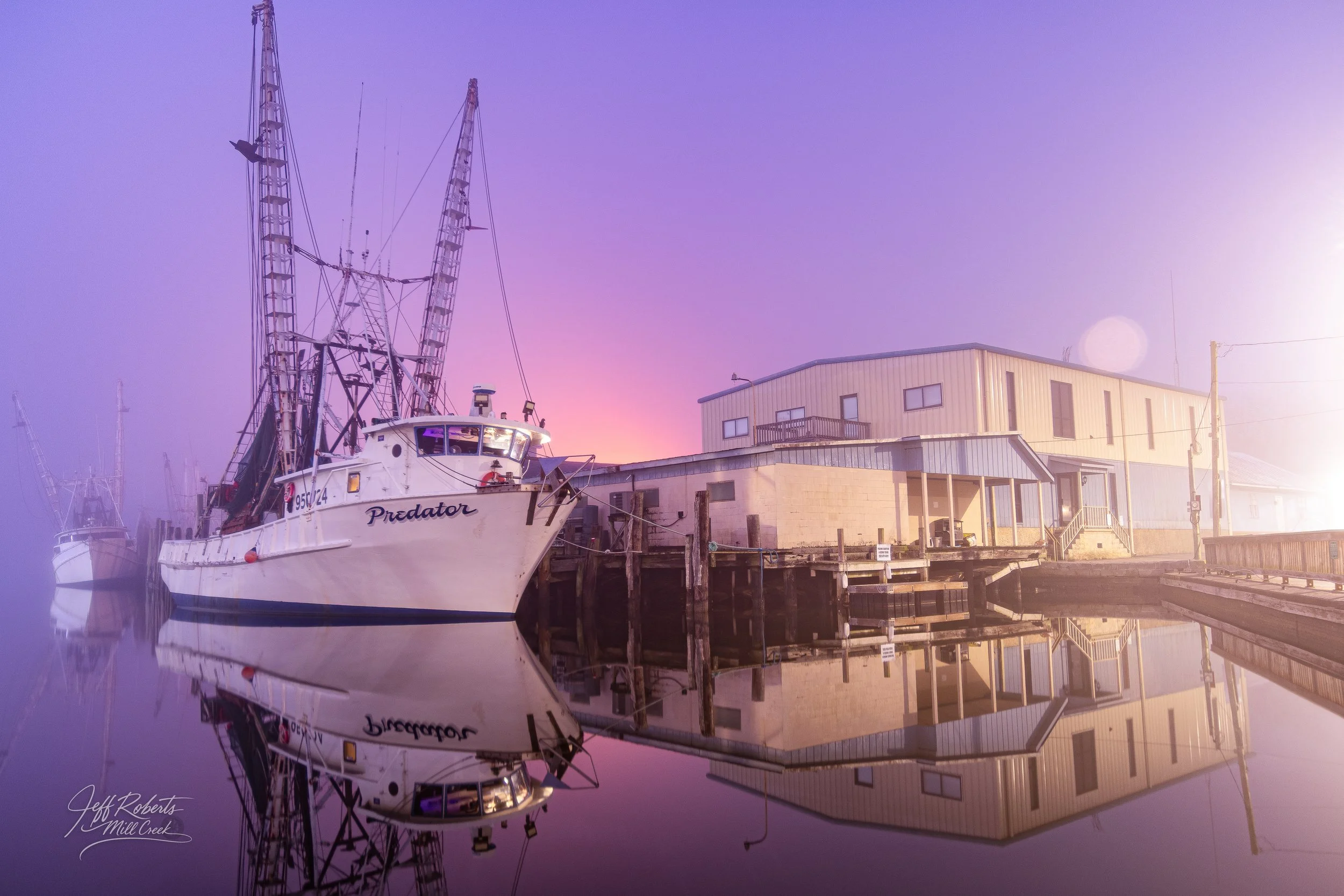 A fishing boat named Predator docked at a pier at sunrise, with buildings in the background and calm water reflecting the boat and surroundings.
