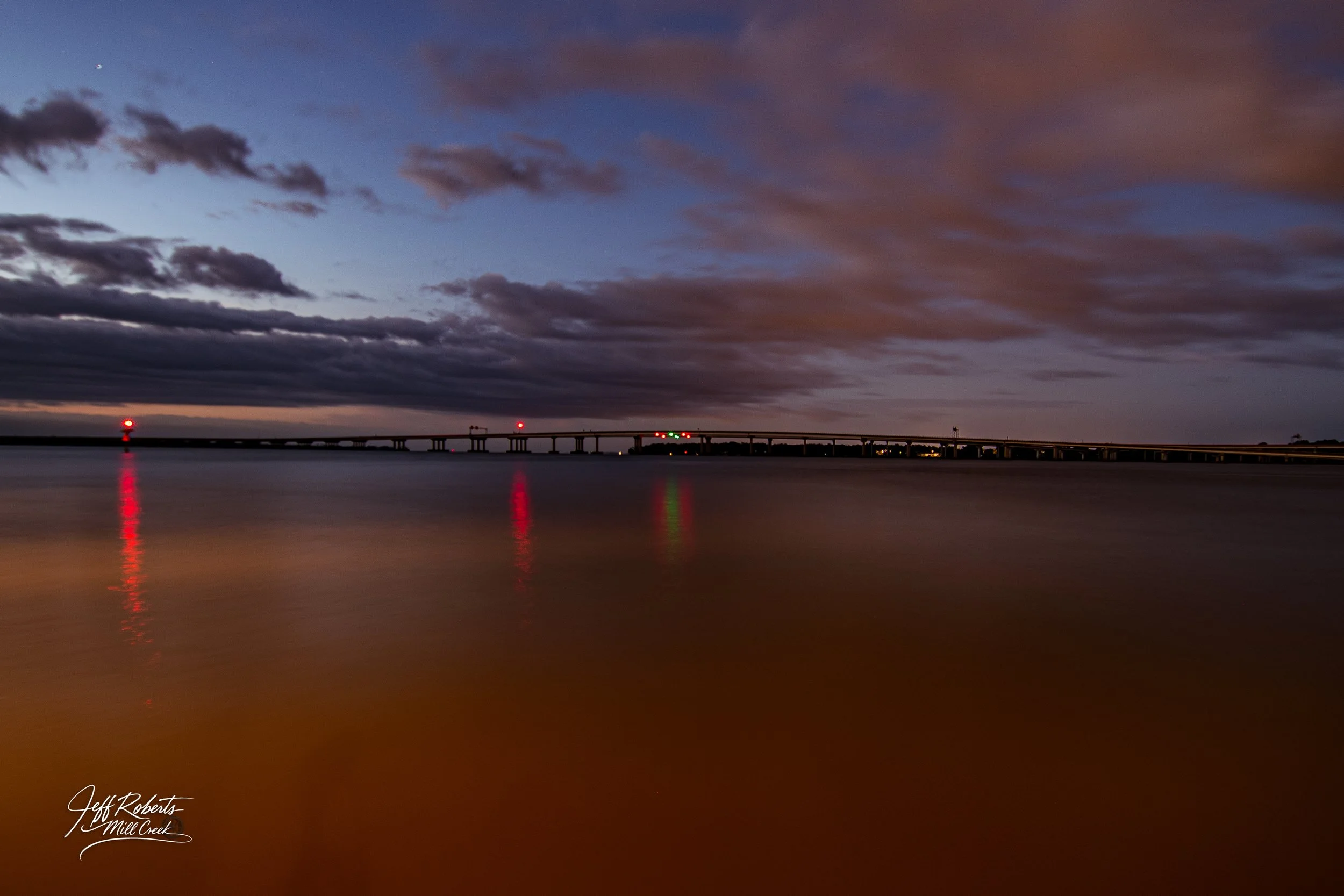 A long bridge at dusk over calm water with reflections of red and green lights, dark clouds in the sky, and a faint star visible on the left.