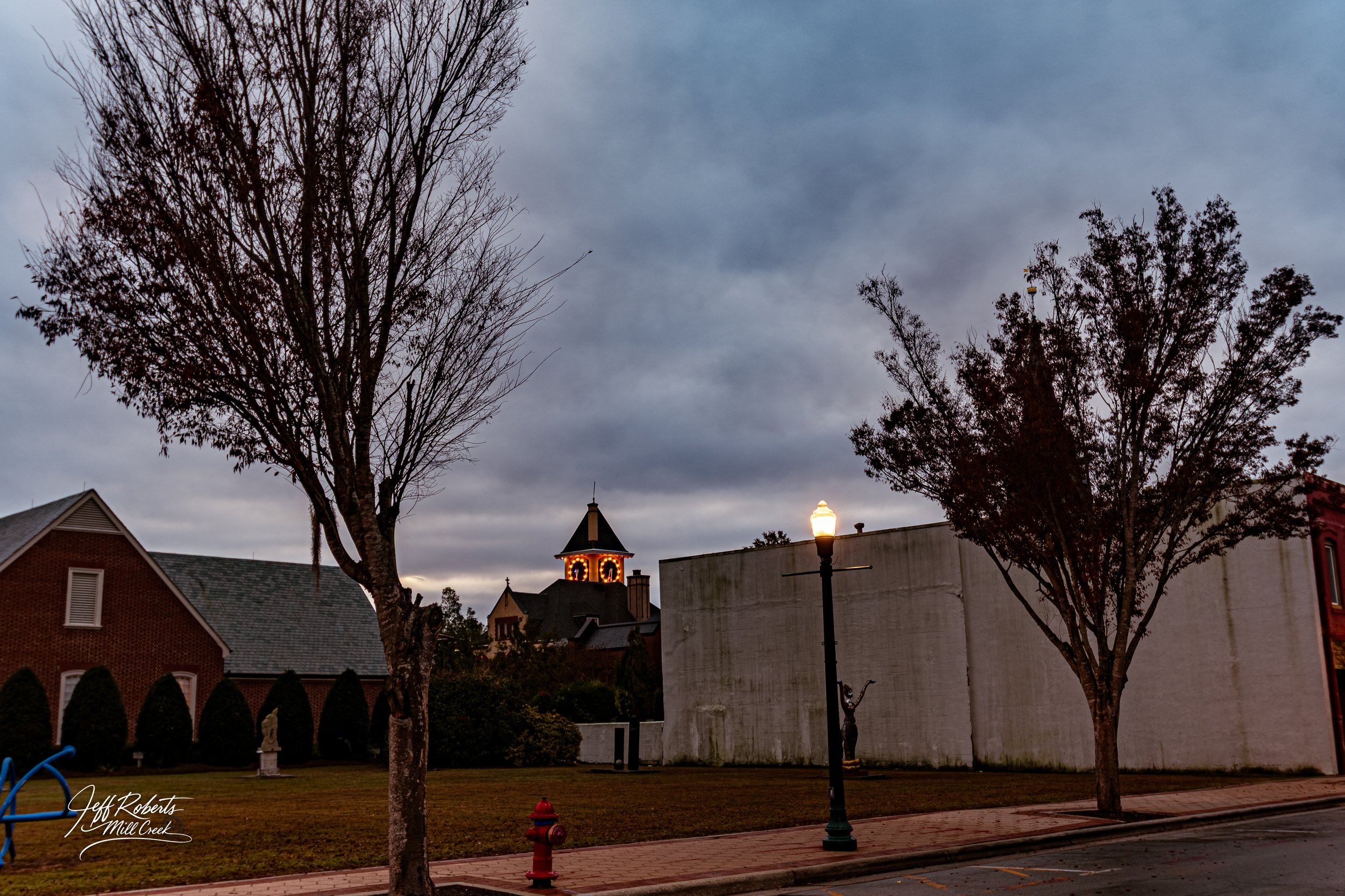 A street scene during dusk with a lit streetlamp, two leafless trees, a brick building, and a church with lit windows and a tall steeple in the background under a cloudy sky.