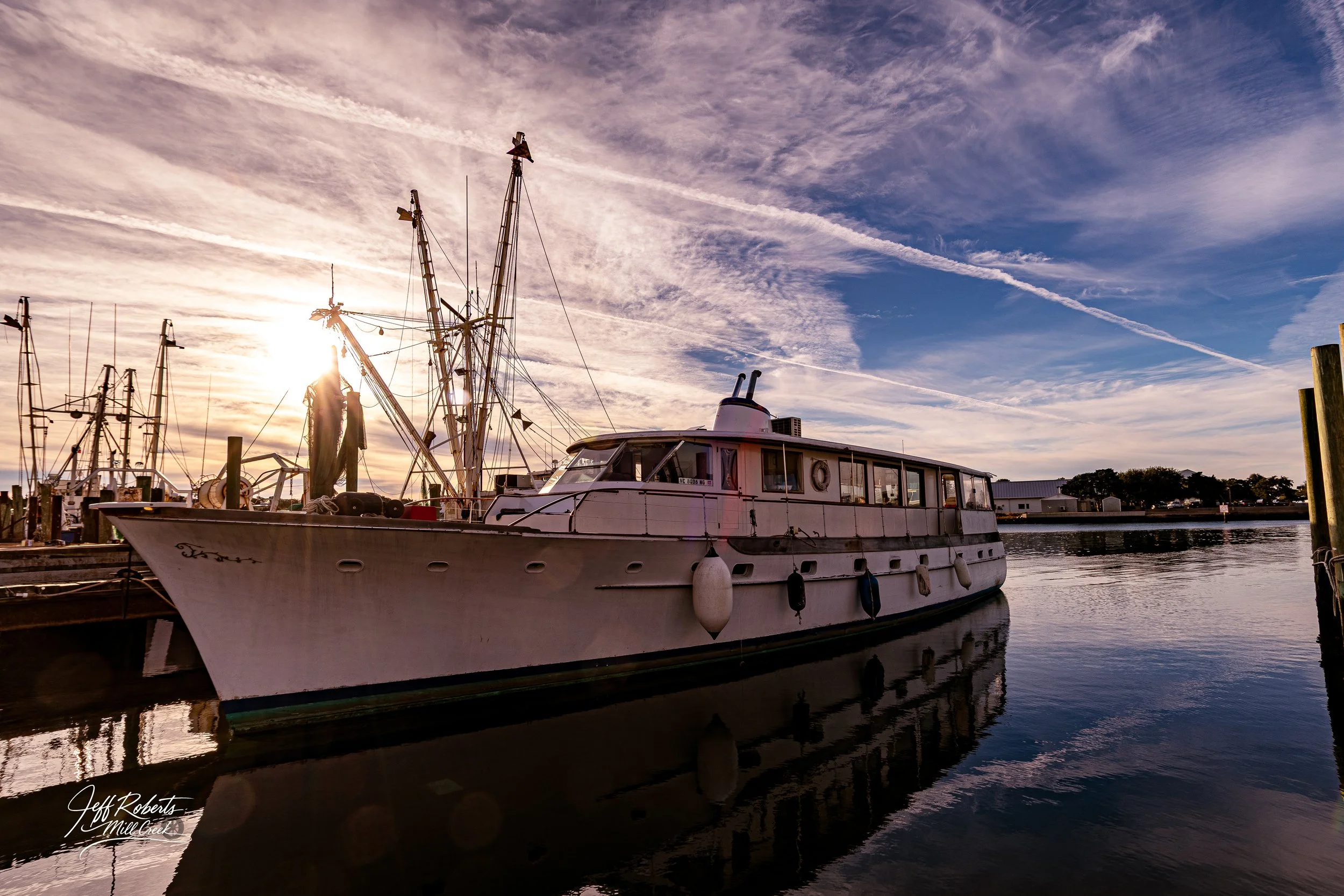 A white boat docked at a marina during sunset, with a partly cloudy sky and streaks of clouds overhead.