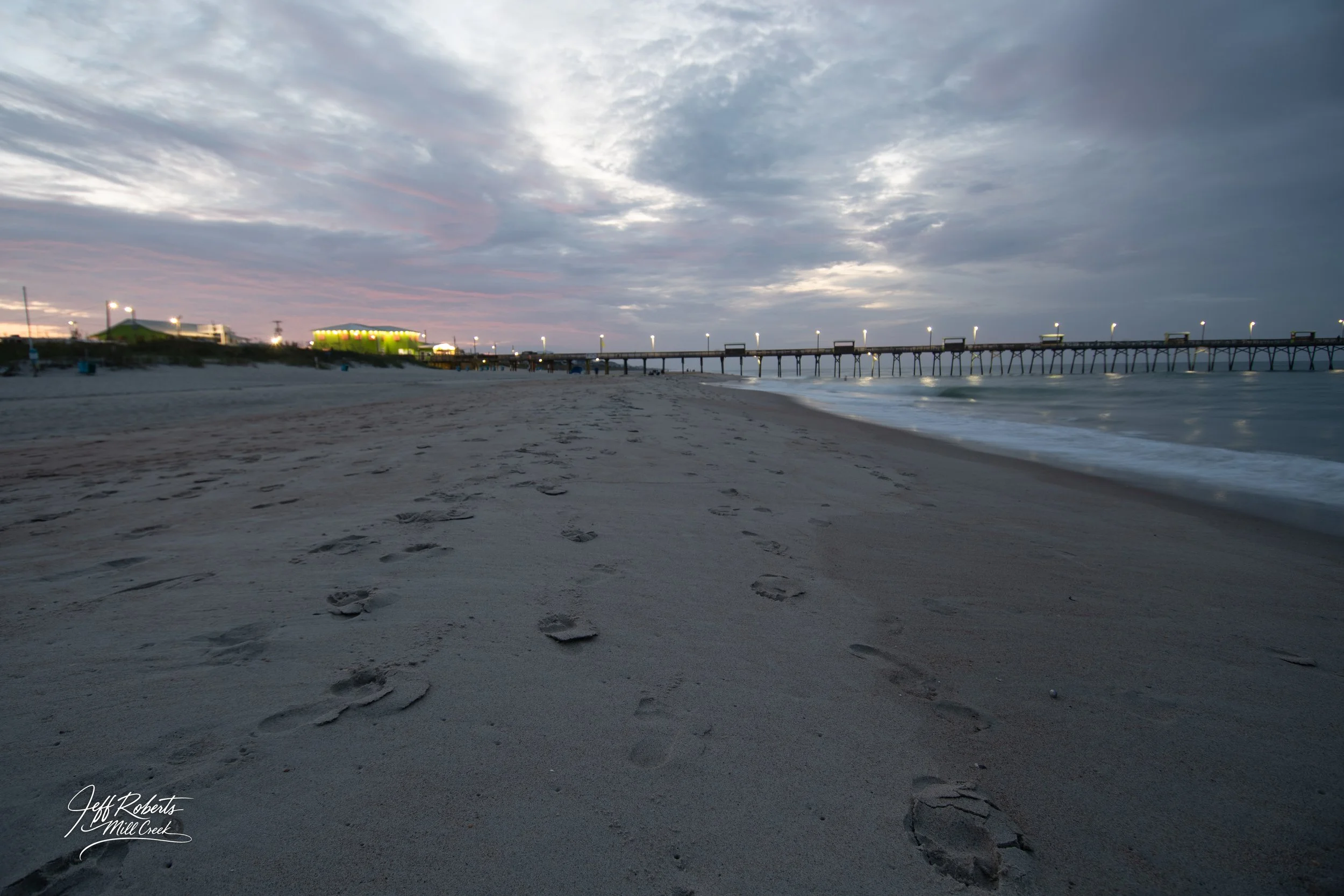 Footprints on a sandy beach near the water with a pier and a building illuminated in the background under cloudy sky during dusk.