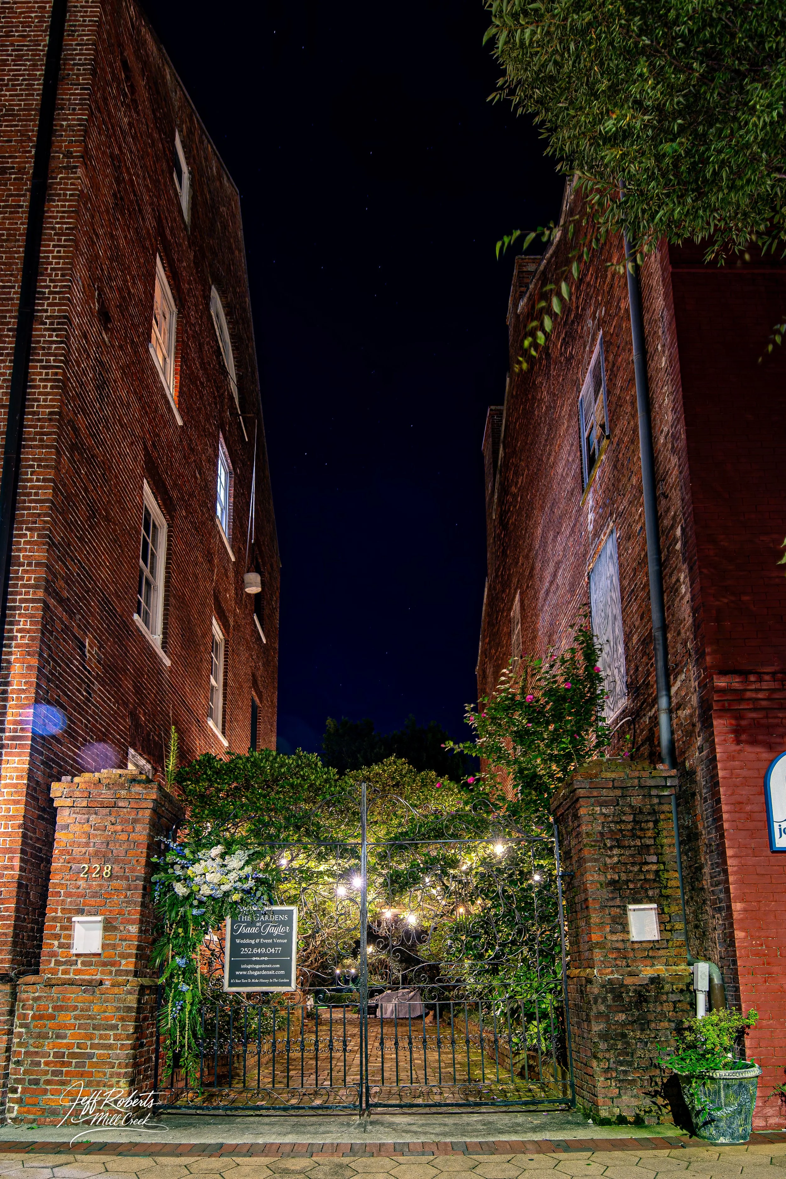 Nighttime view of a brick alleyway between two tall brick buildings, with a wrought iron gate decorated with floral arrangements and string lights leading into a lush garden area.