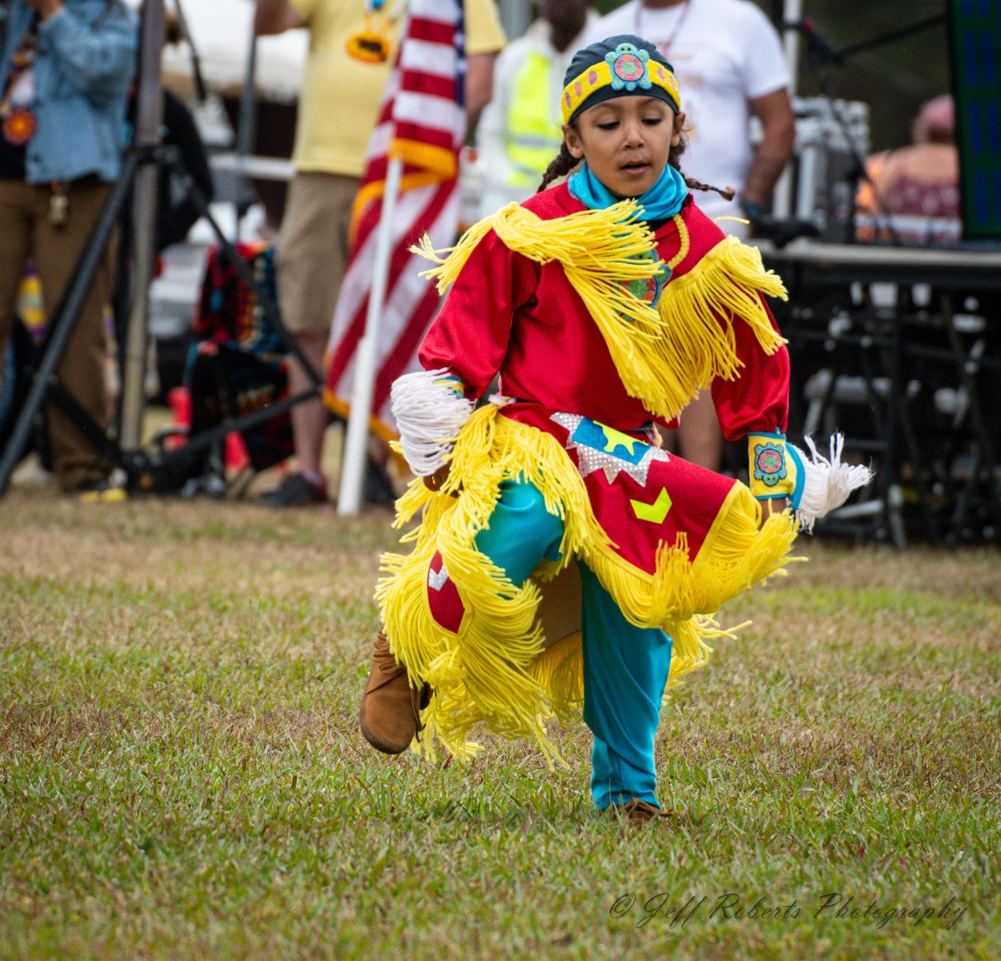 Young girl in colorful traditional Native American dance regalia performing at cultural event on grassy field, with spectators and equipment visible in background.