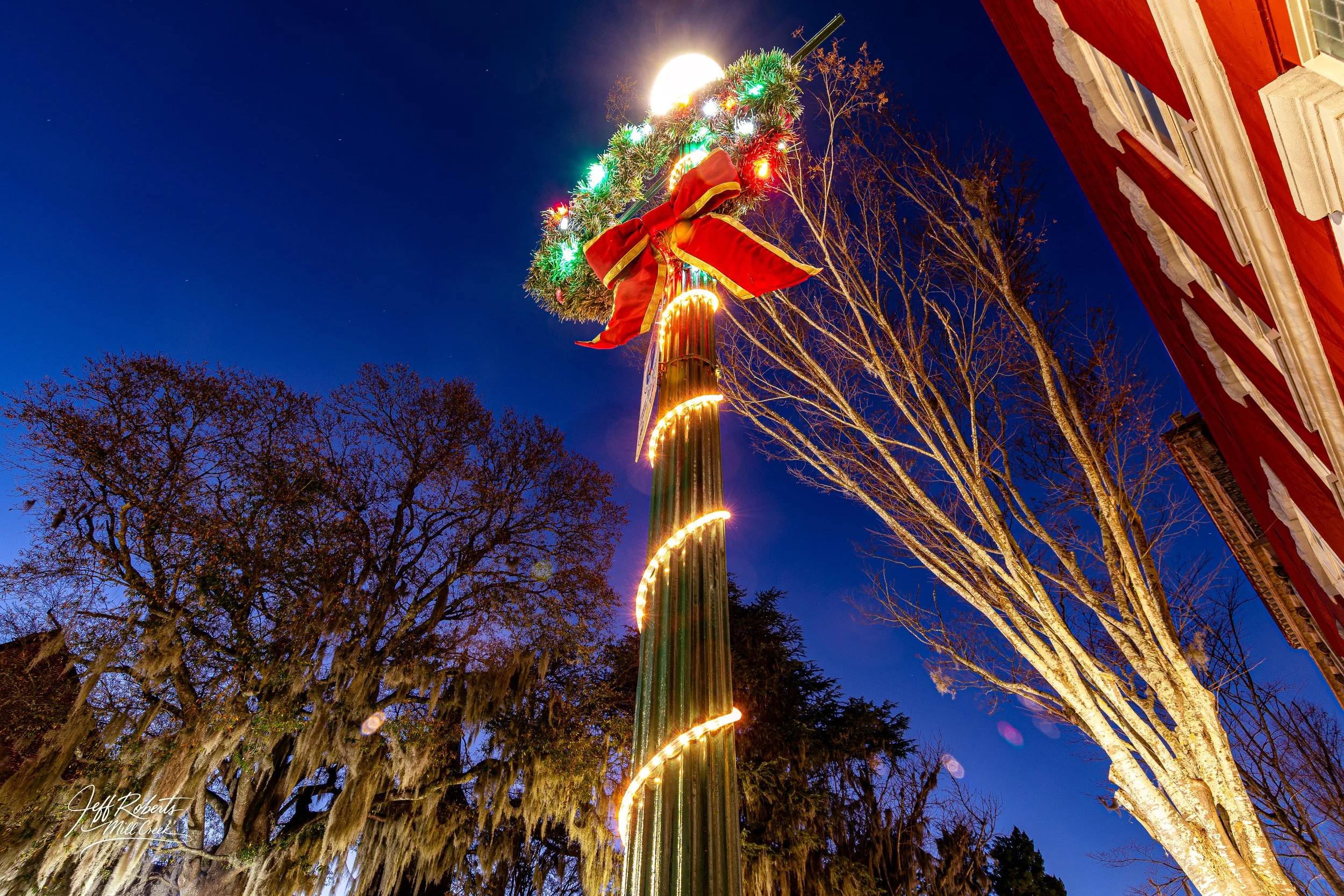 A decorated street pole topped with a wreath, a red bow, and Christmas lights at night, with trees and buildings in the background.