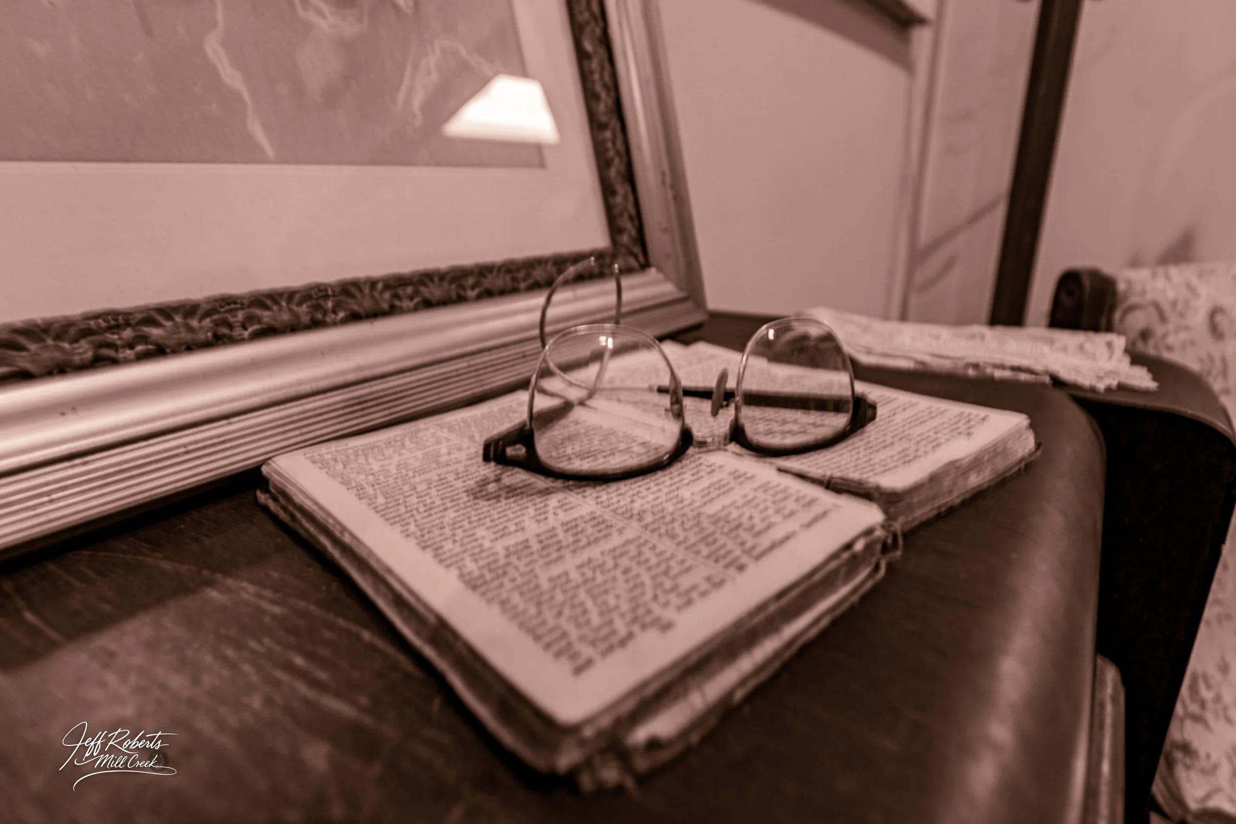 A pair of eyeglasses resting on an open Bible on a wooden surface with framed artwork and decorative items in the background.