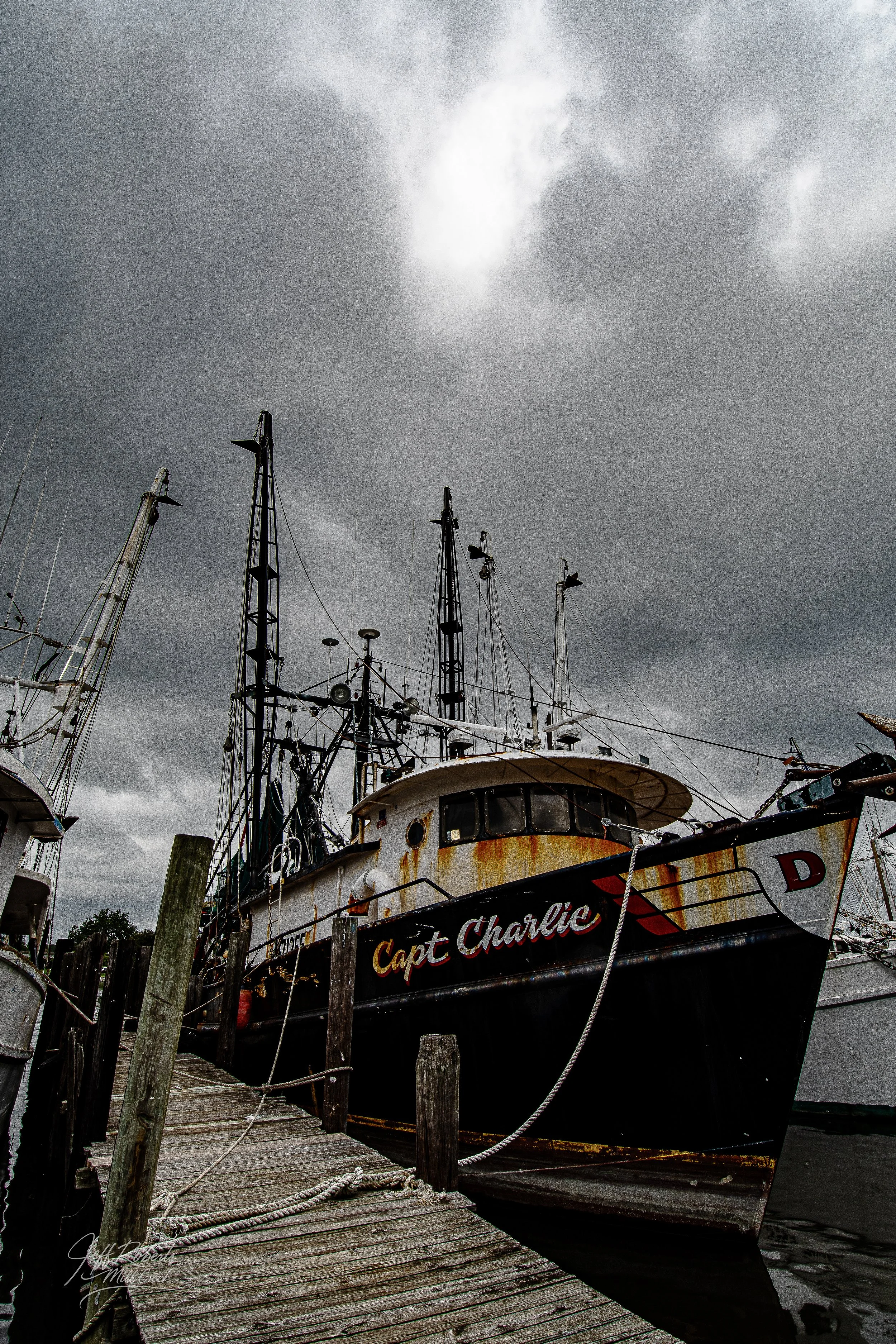 A rusty, black fishing boat named 'Capt Charlie' docked at a weathered wooden pier under a dark, cloudy sky.