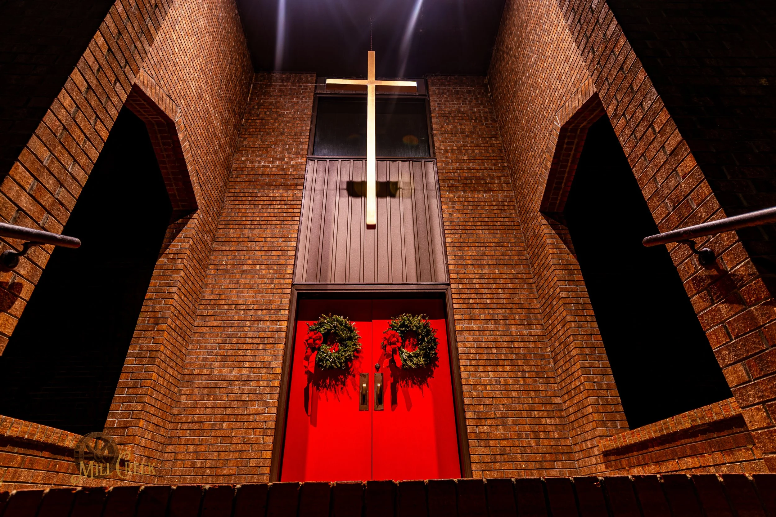 Bright red church doors decorated with two Christmas wreaths and a large wooden cross mounted on the brick facade above the entrance at night.