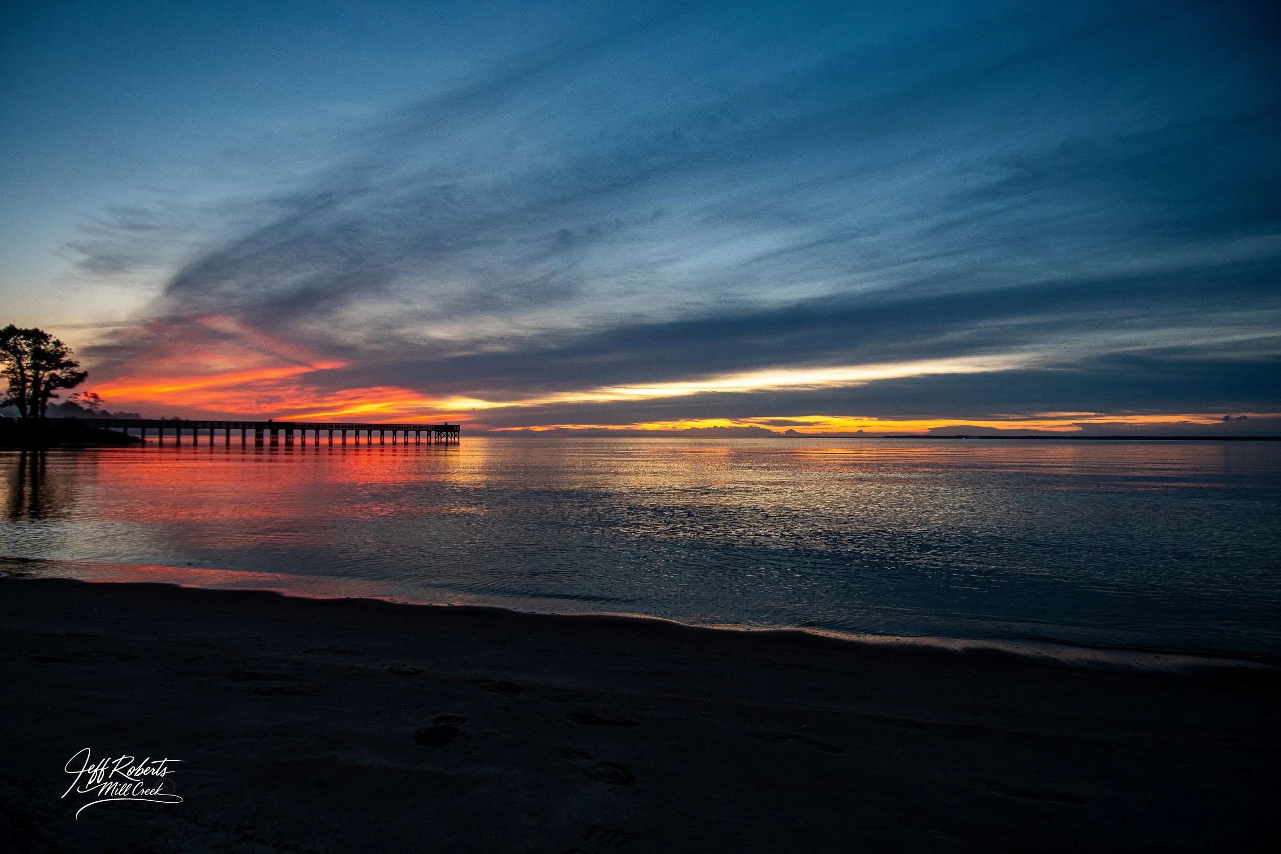Sunset over a calm body of water with a pier extending into the distance on the left, and a few trees silhouetted on the shoreline. Bright orange and yellow hues are visible near the horizon with dark clouds overhead.