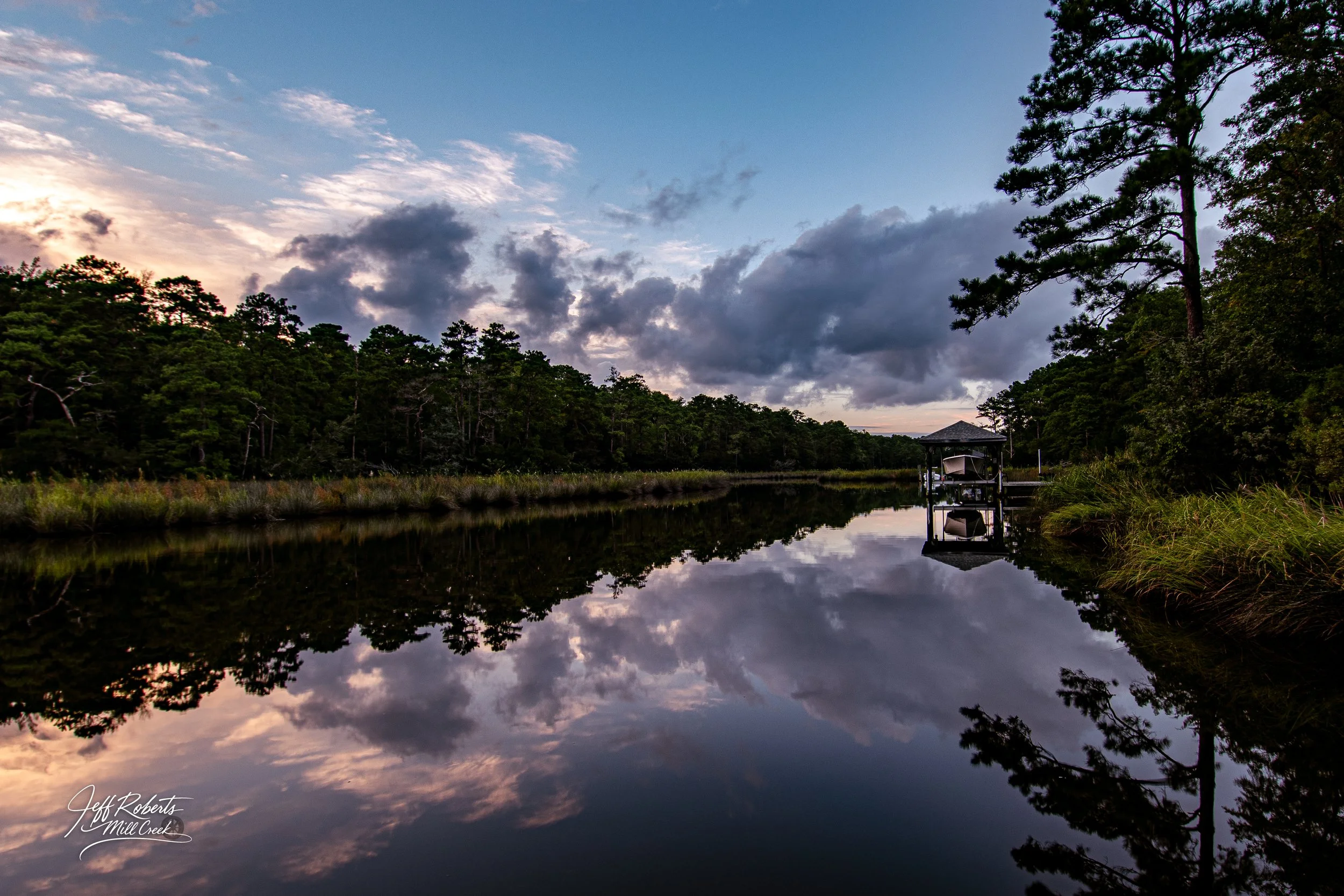 Serene river scene at dusk with calm water reflecting the cloudy sky, trees on both sides, and a small dock with a boat on the right.