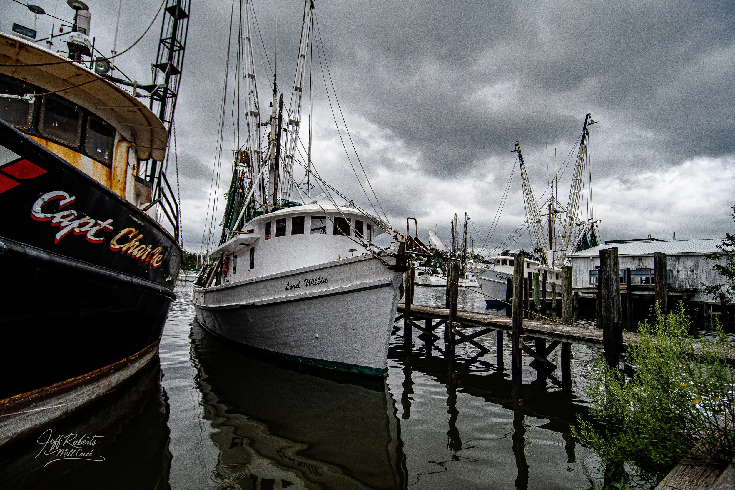 Several boats docked at a marina on a cloudy day, including a white boat named Lord Willin and other boats in the background. The scene features a wooden pier and stormy skies.