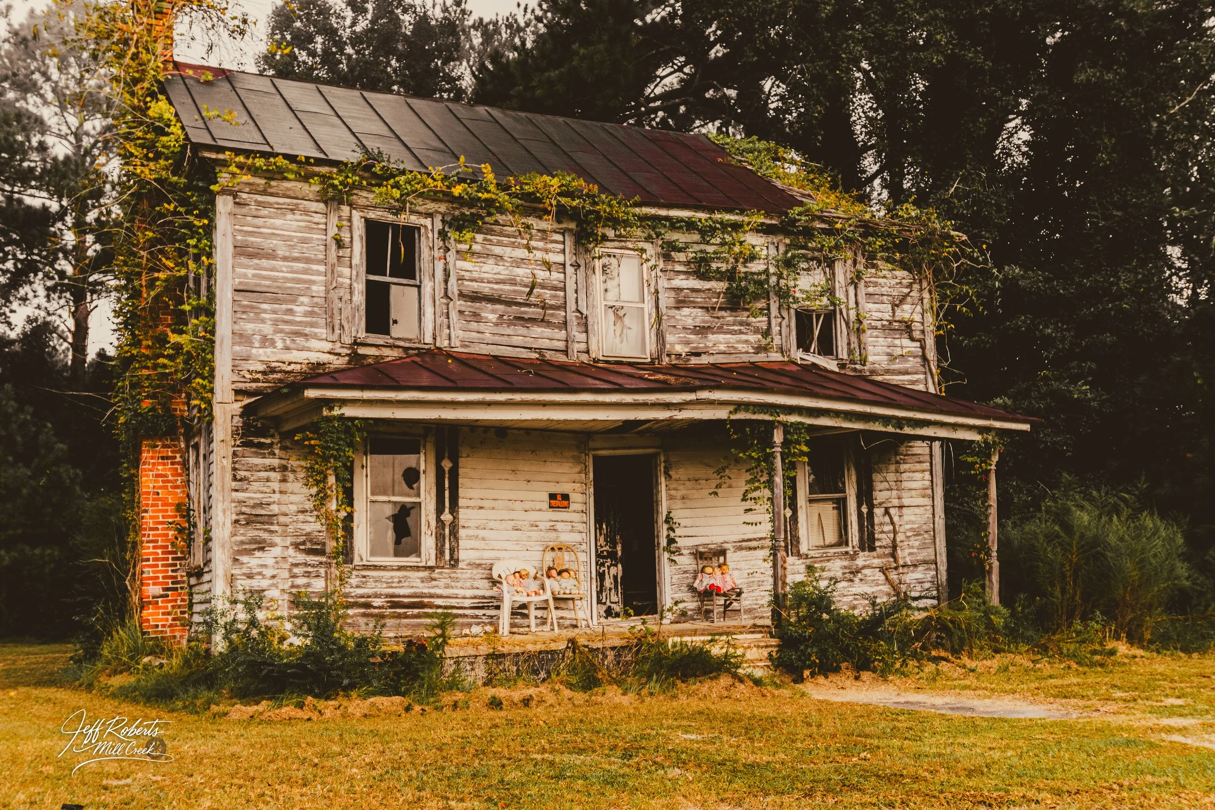 An old, abandoned two-story house with peeling white paint, broken windows, and overgrown vines on the exterior. The front porch has three chairs with stuffed animals, and the surrounding yard is unkempt with tall grass and shrubs. Trees are visible 
