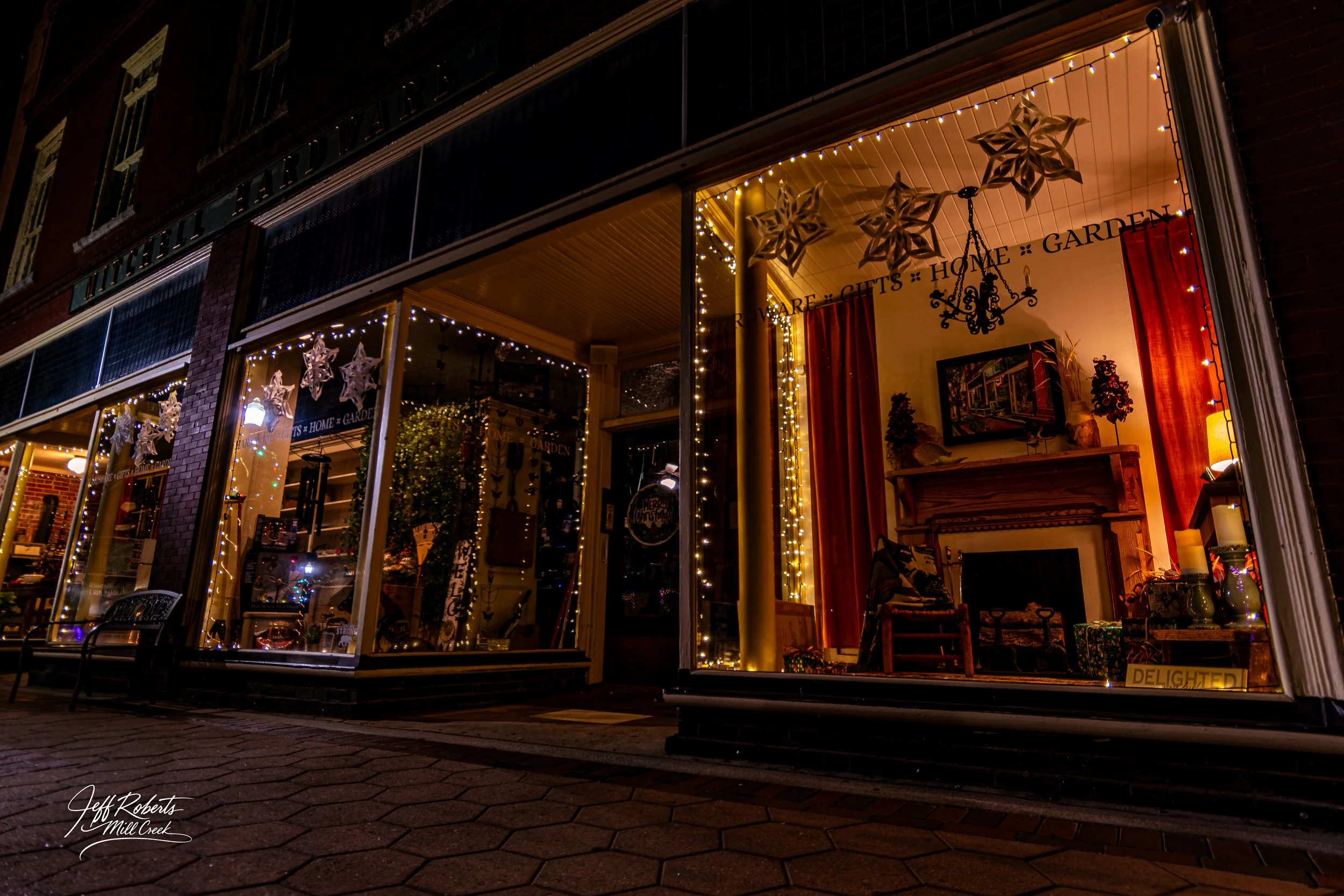 Nighttime view of the exterior of a gift shop decorated with string lights and hanging star ornaments, with visible items inside on display through large windows.