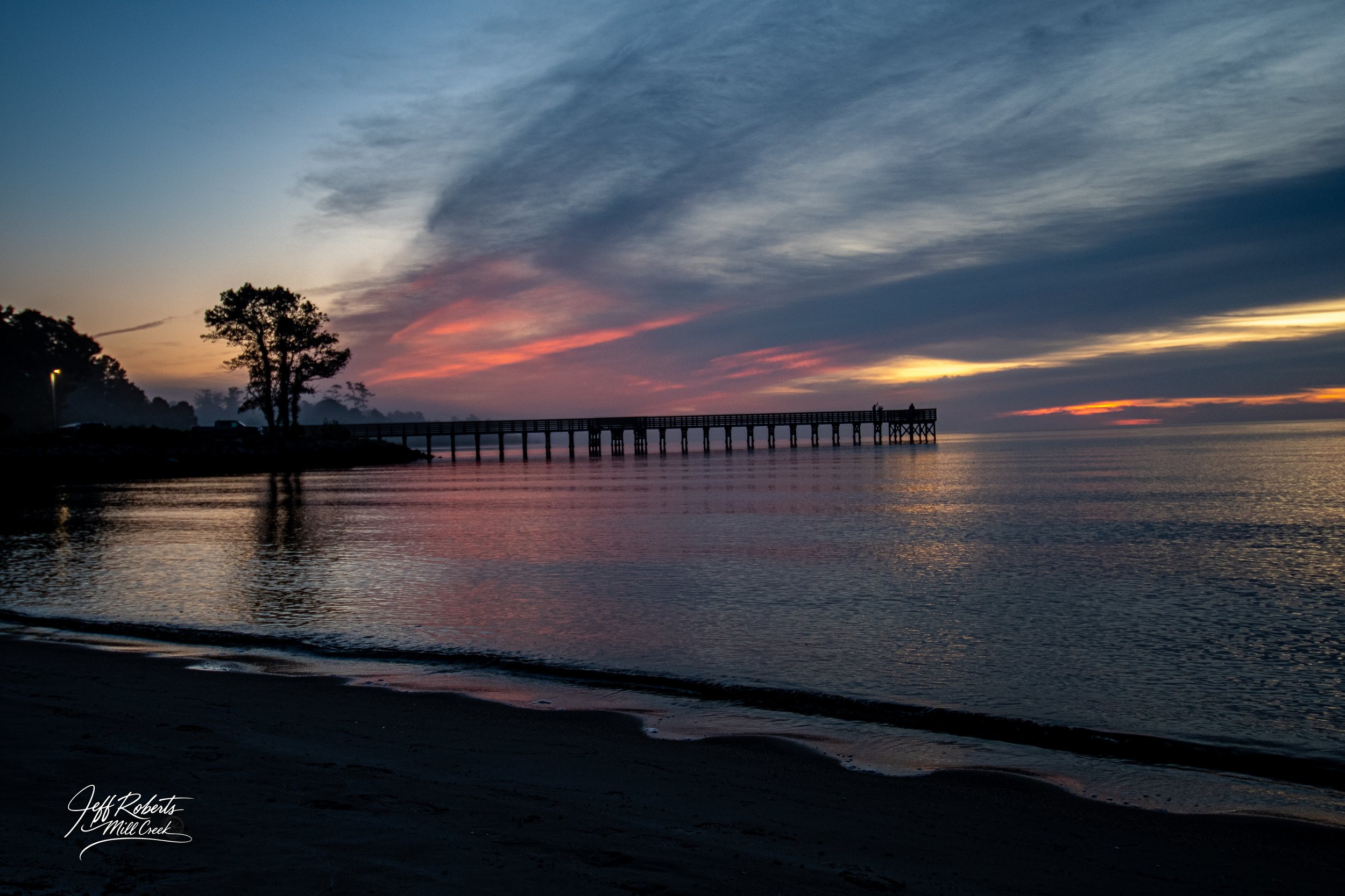 A sunset over a calm body of water with a pier extending out into the water and trees on the land.