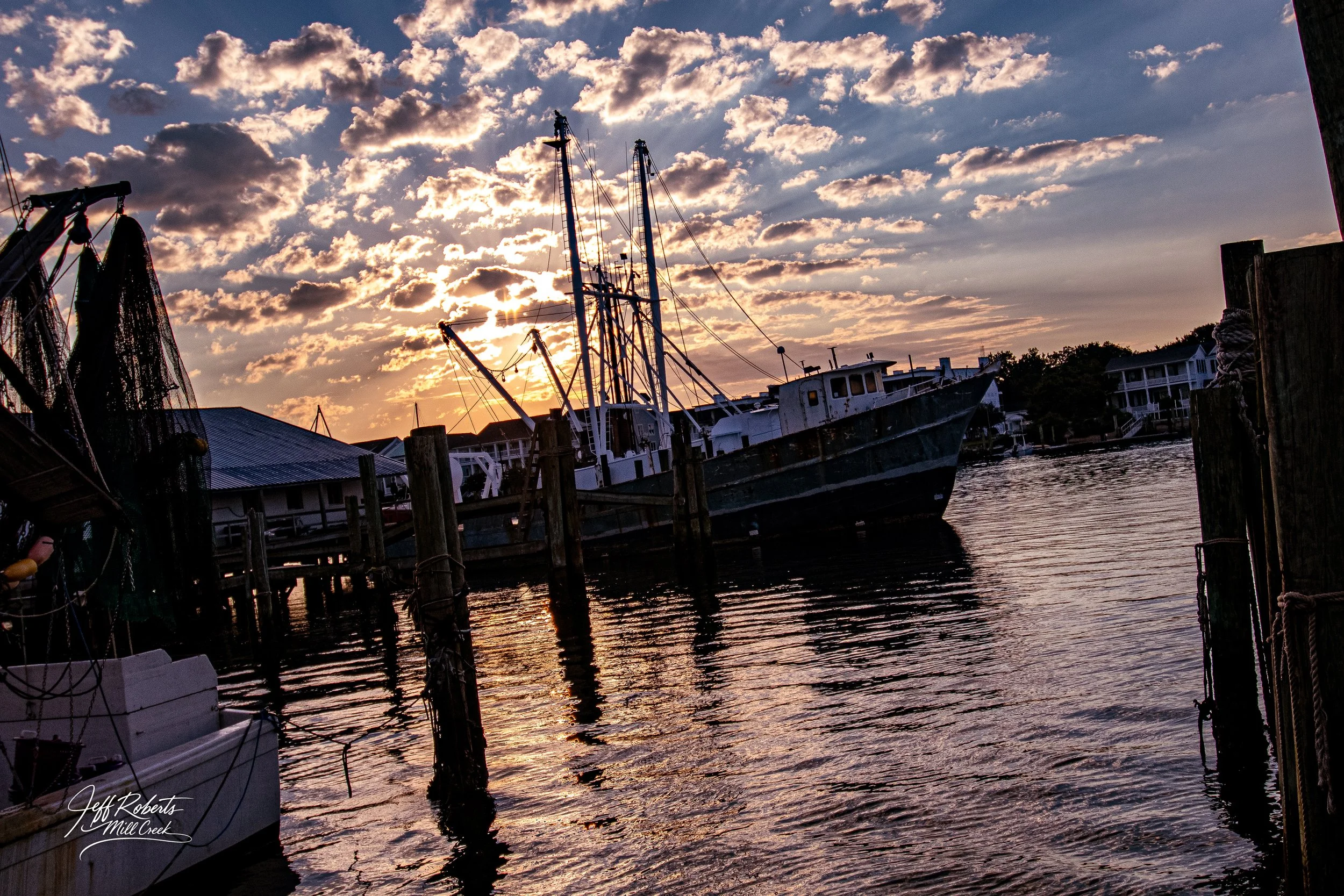A boat docked at a marina during sunset, with clouds in the sky and buildings in the background.