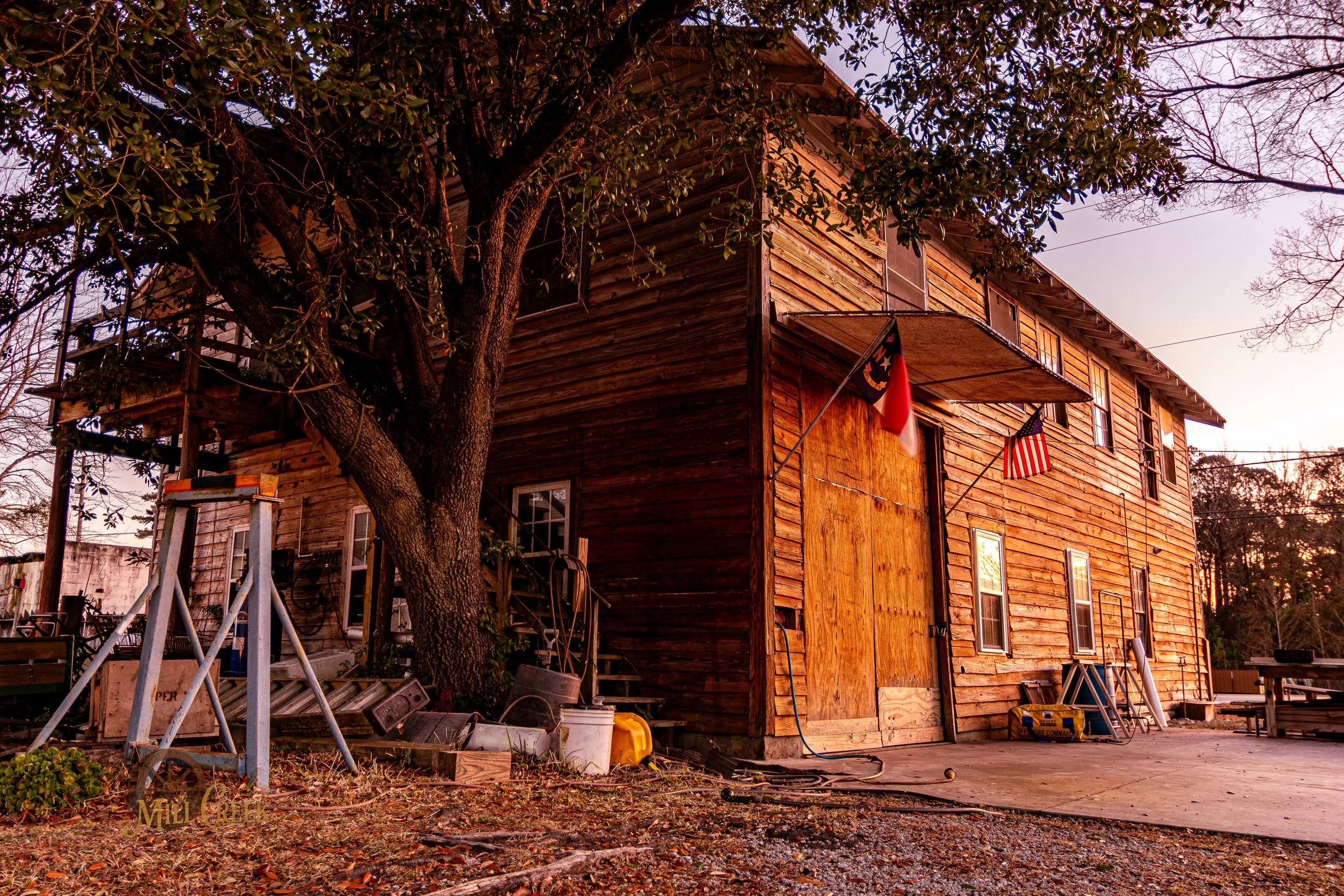 A two-story wooden house with an American flag and a black, red, and yellow flag hanging from the front. There is a large tree next to the house with a swing set and various tools and containers scattered around.