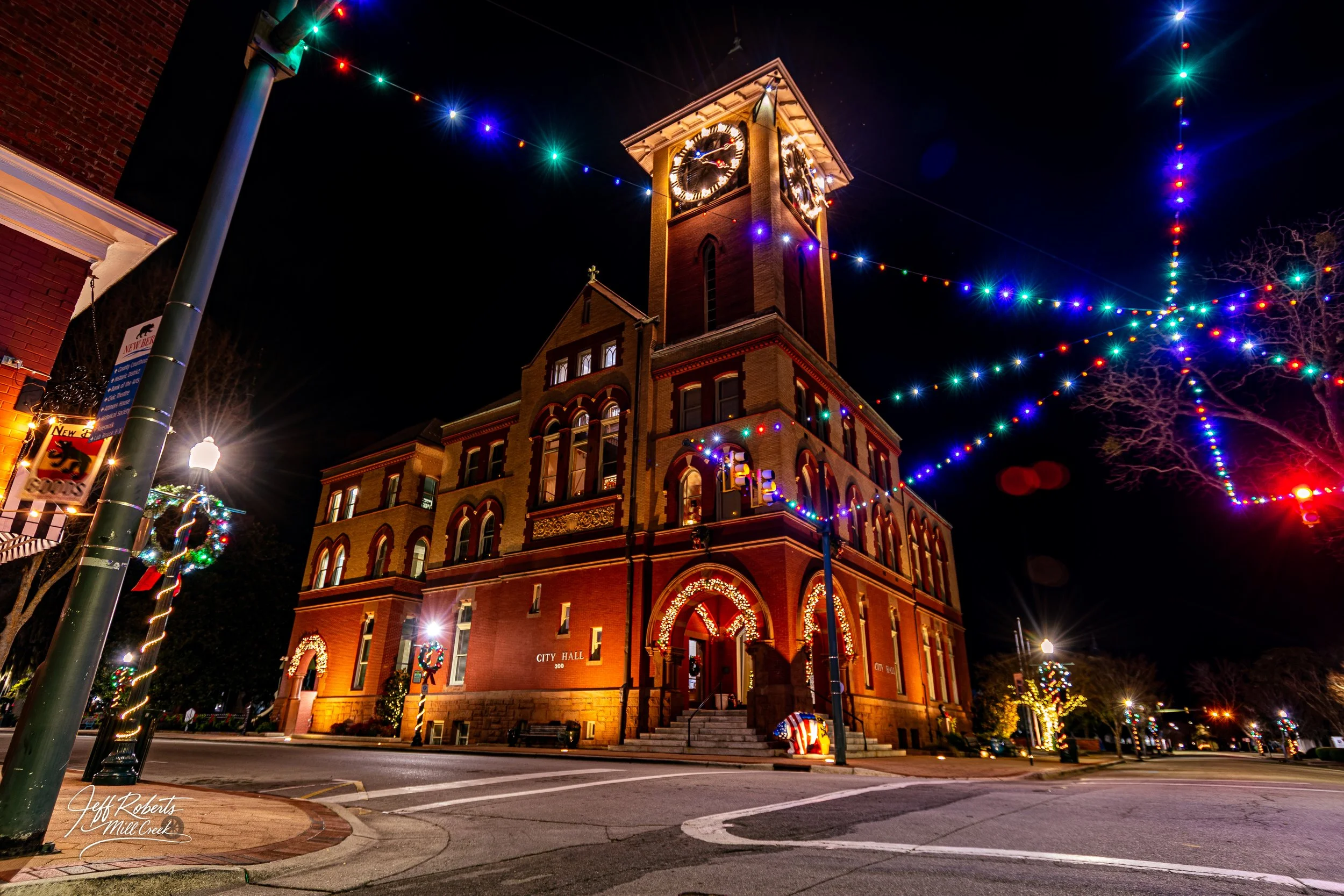 A historic city hall building decorated with Christmas lights and wreaths, with colorful string lights hanging across the street at night.