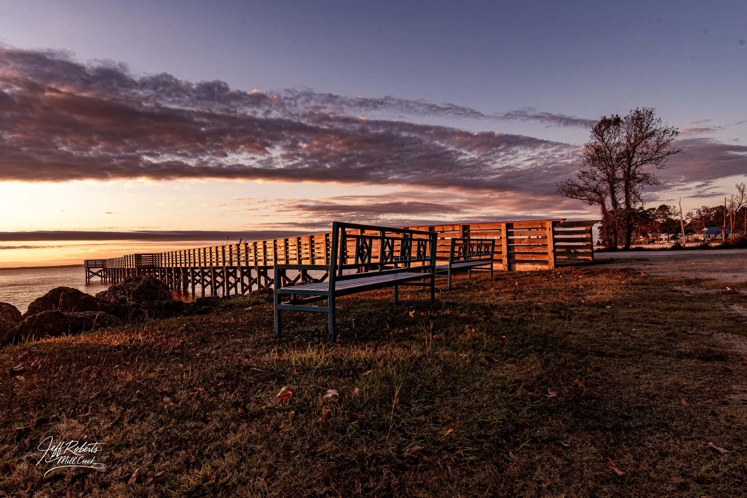 Sunset over a pier with benches and rocks, with clouds in the sky and a few trees in the background.