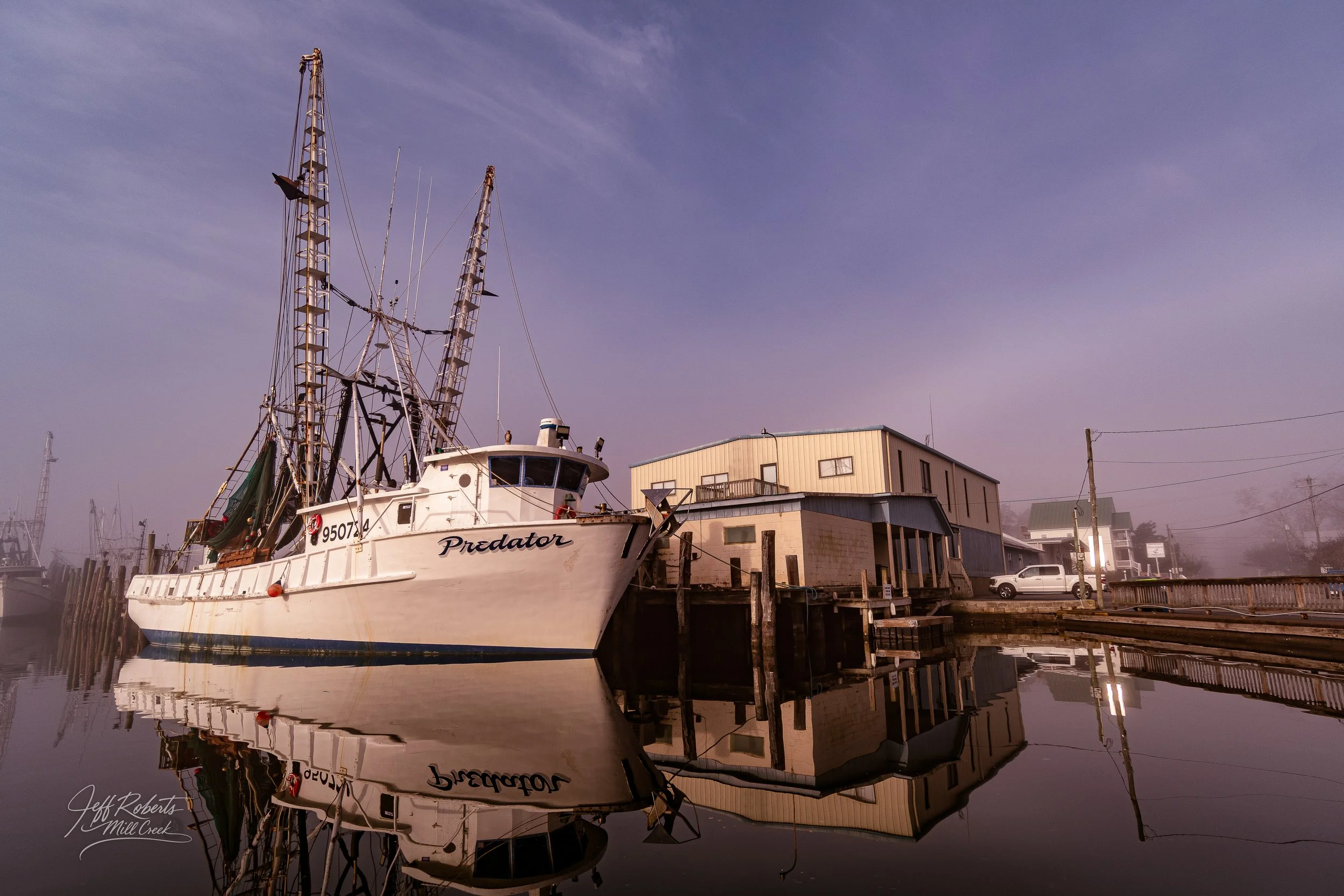 A white fishing boat named Predator docked at a marina, with calm water reflecting the boat and surrounding buildings, under a foggy sky.