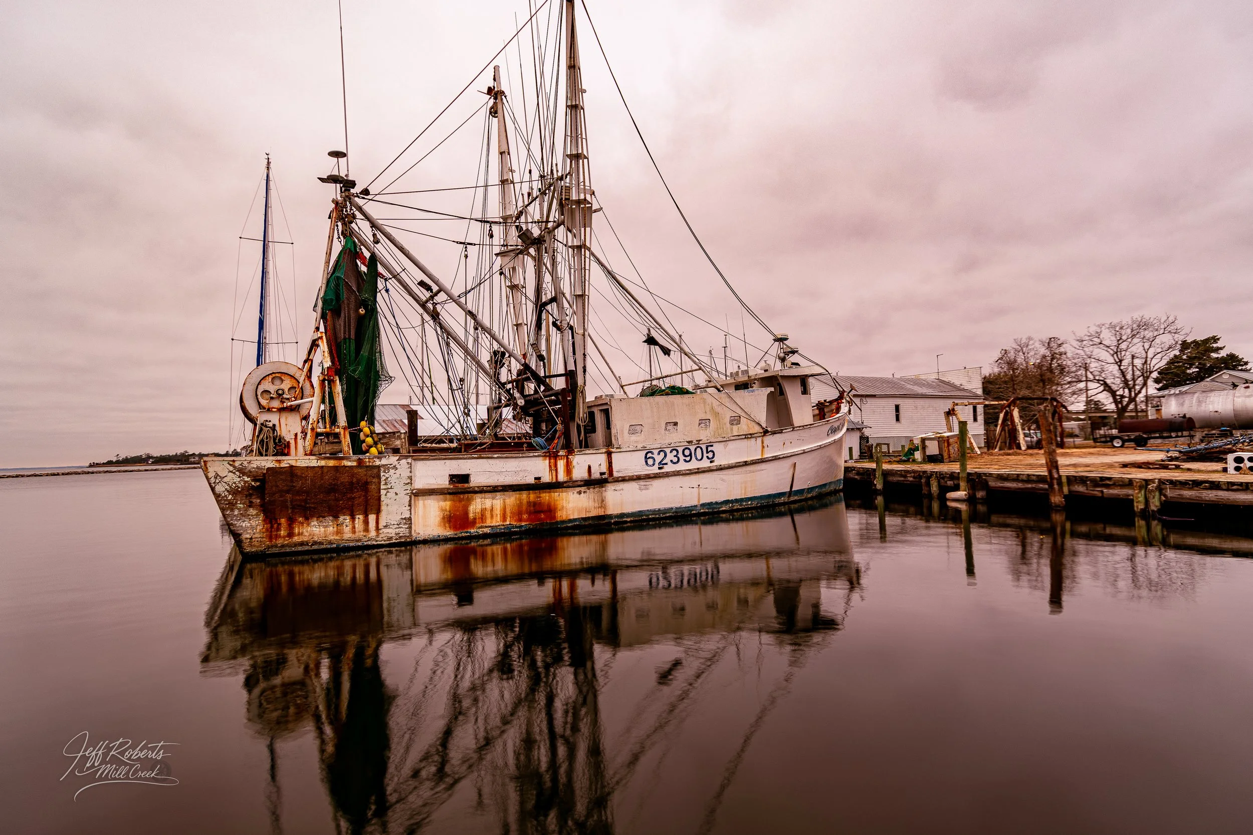 Old rusted fishing boat docked at a pier with calm water reflecting it, overcast sky, and small buildings and trees in background.