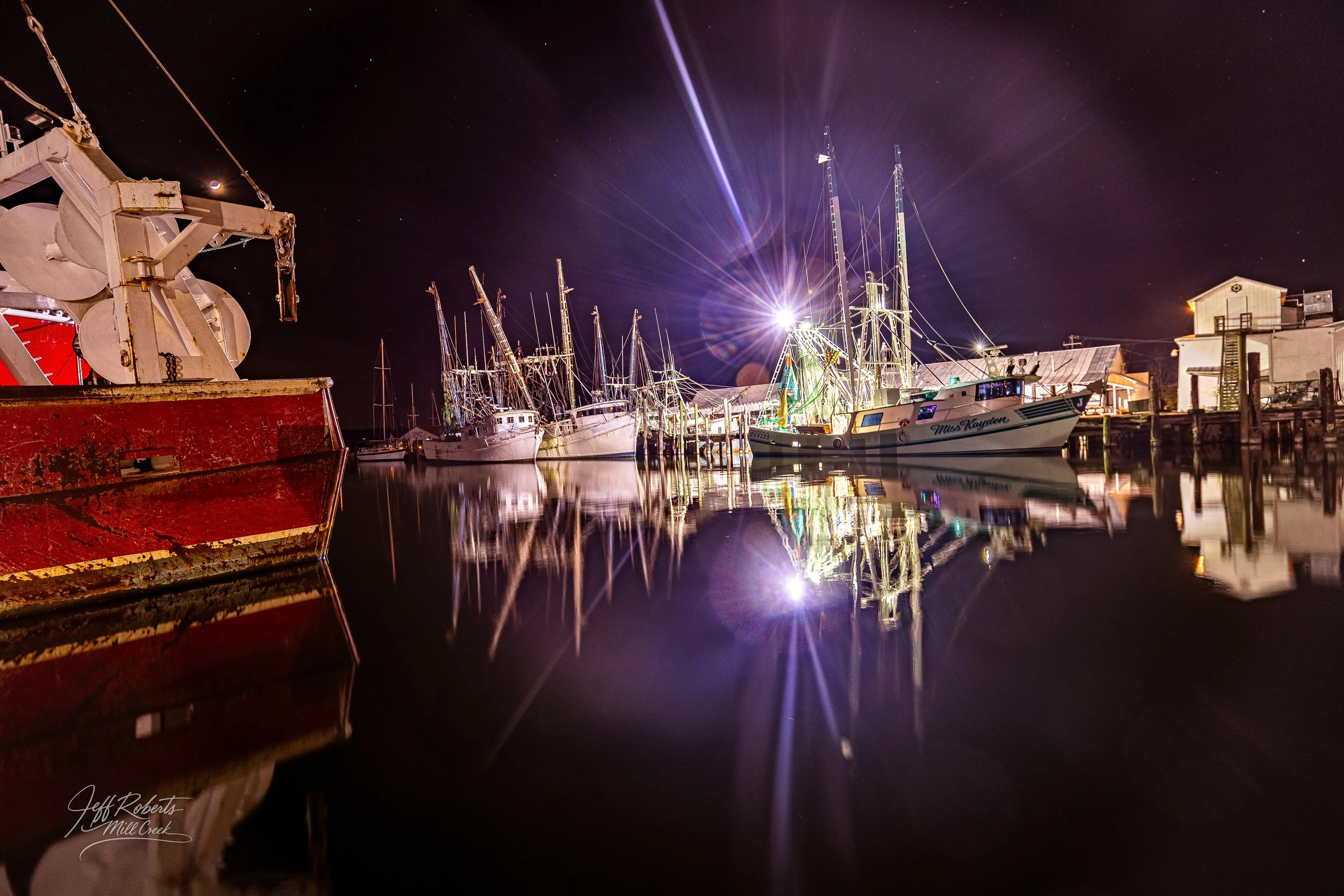 Nighttime view of boats docked at a marina with calm water reflecting their images and a bright light illuminating the scene, along with a clear night sky with stars.