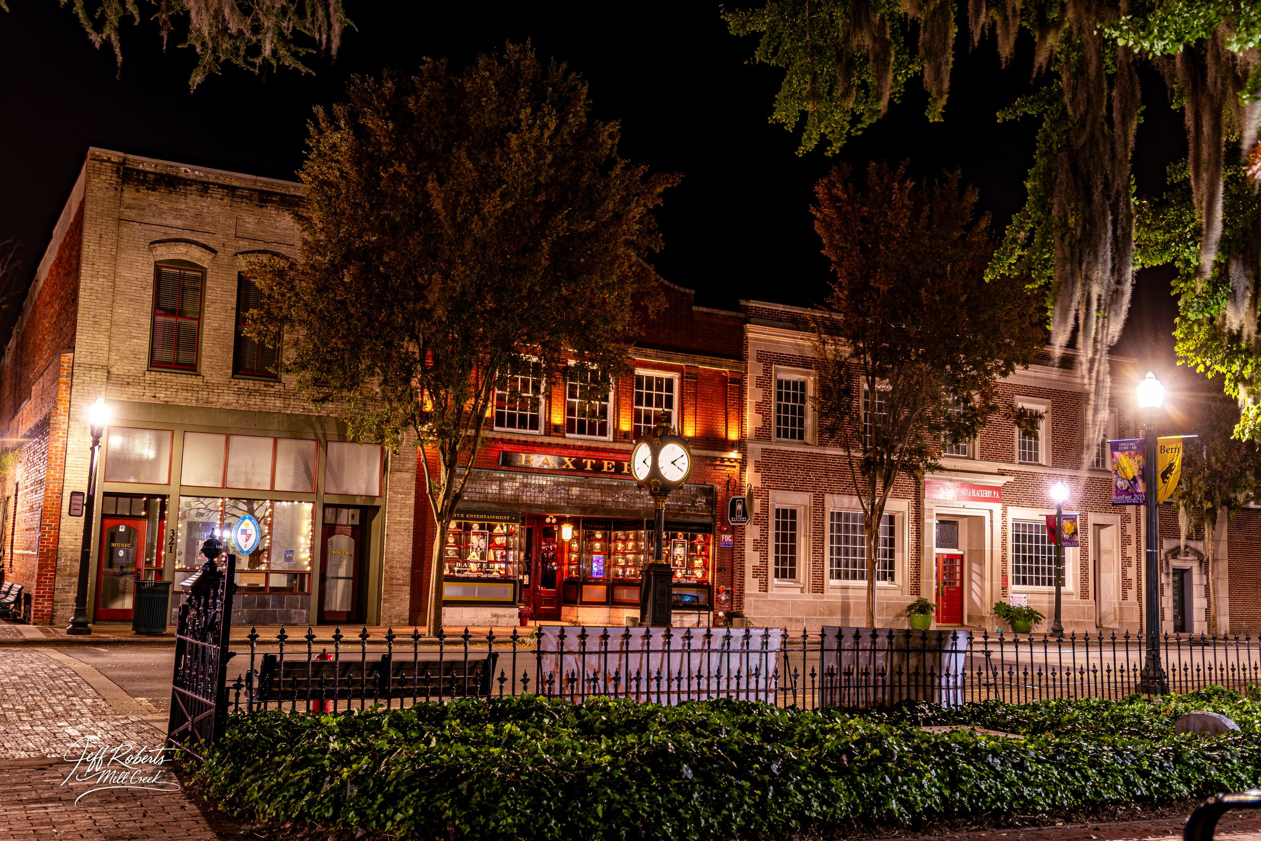 Night view of a historic downtown street with brick buildings, street lamps, a clock, trees, and storefronts illuminated at night.