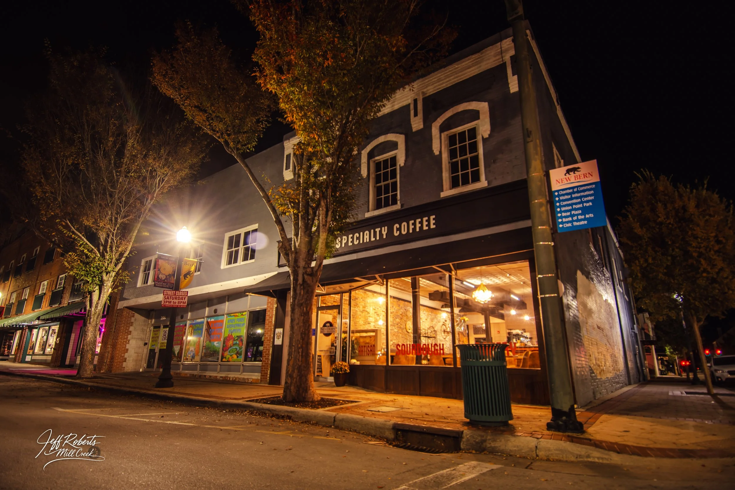 Nighttime view of a corner coffee shop with large glass windows, warm interior lighting, and a sign that says 'Specialty Coffee.' The building is painted black with white accents and are two stories tall. There are trees with autumn leaves and street