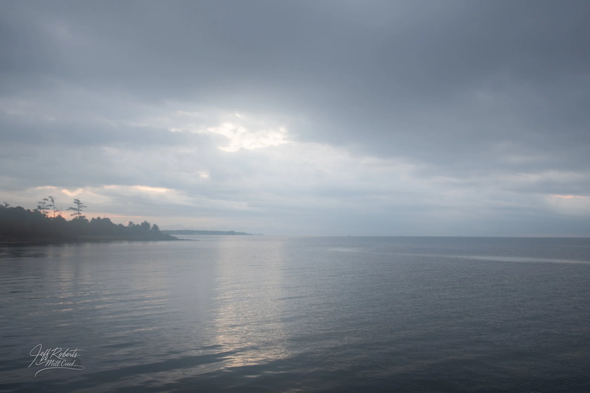 A calm body of water under a cloudy sky with some light breaking through, and a tree-lined shoreline on the left.