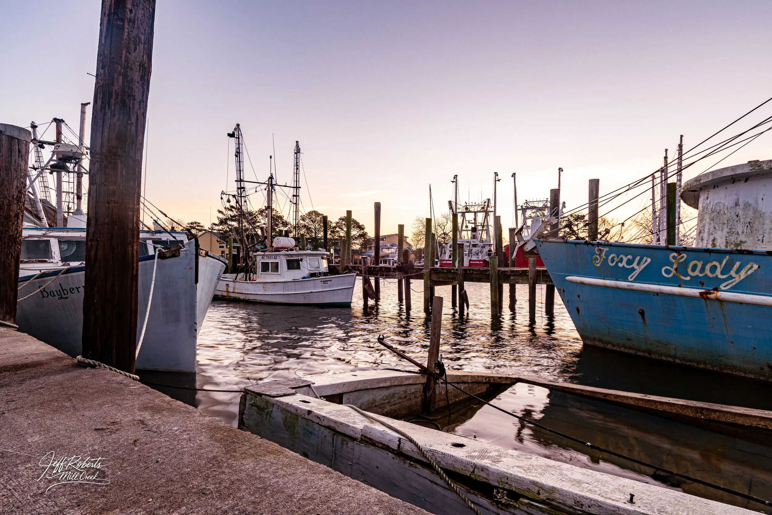 Fishermen's boats docked at a marina during sunset, with wooden posts and calm water in the scene.
