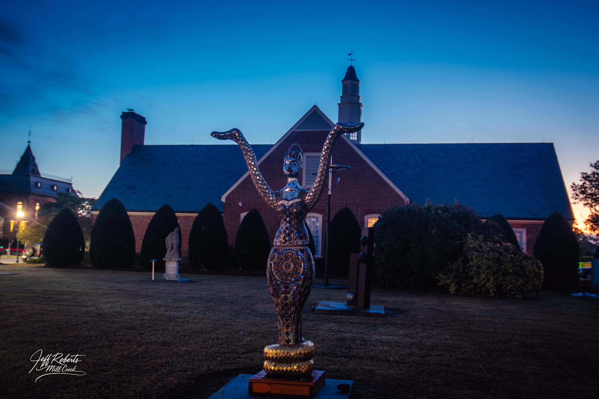 Sculpture of a woman with arms raised, made of reflective, mosaic-like material, in front of a brick church at dusk.