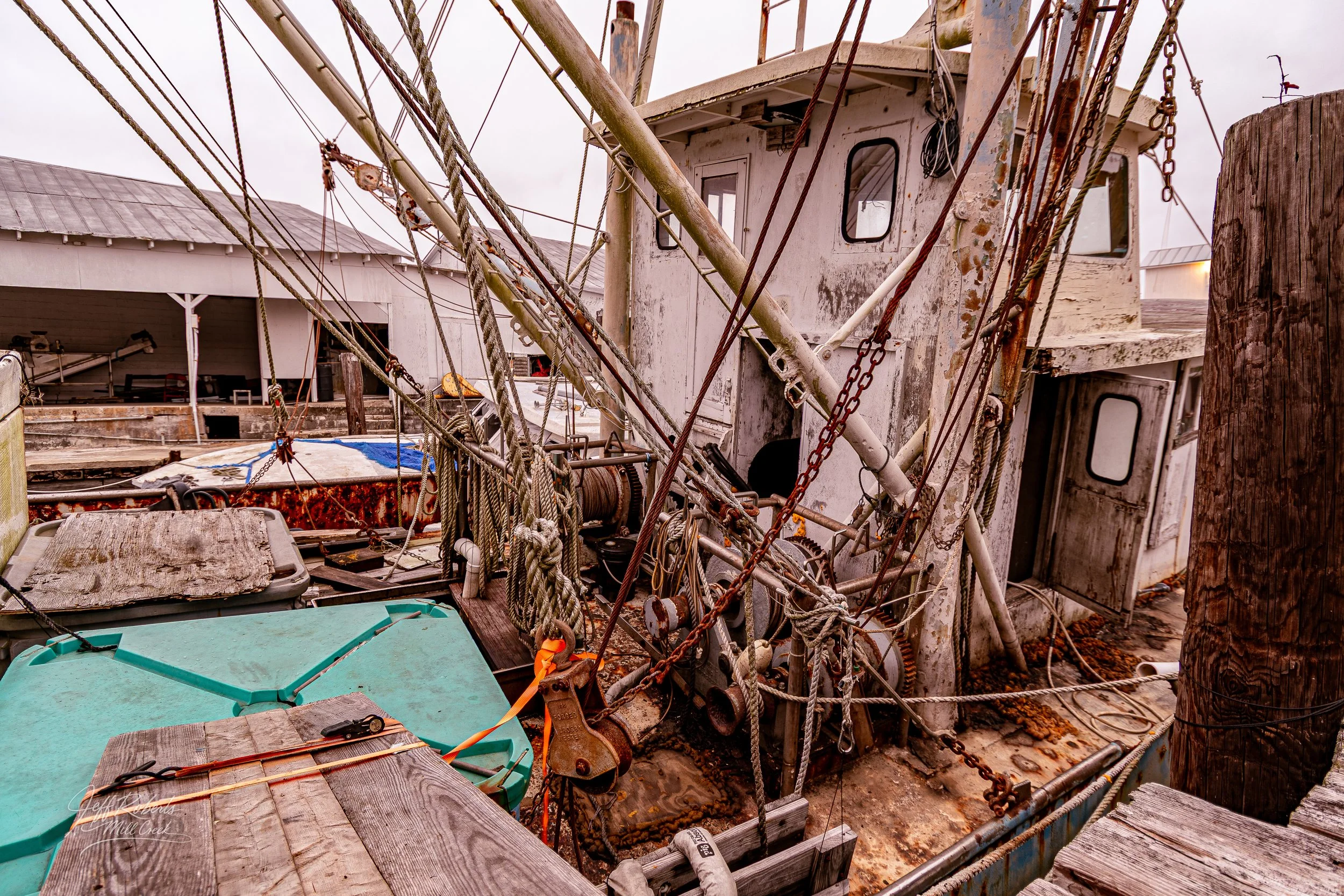 Old, rusty fishing boat with tangled ropes, chains, and equipment in a dockyard, showing weathered wood and metal surfaces.