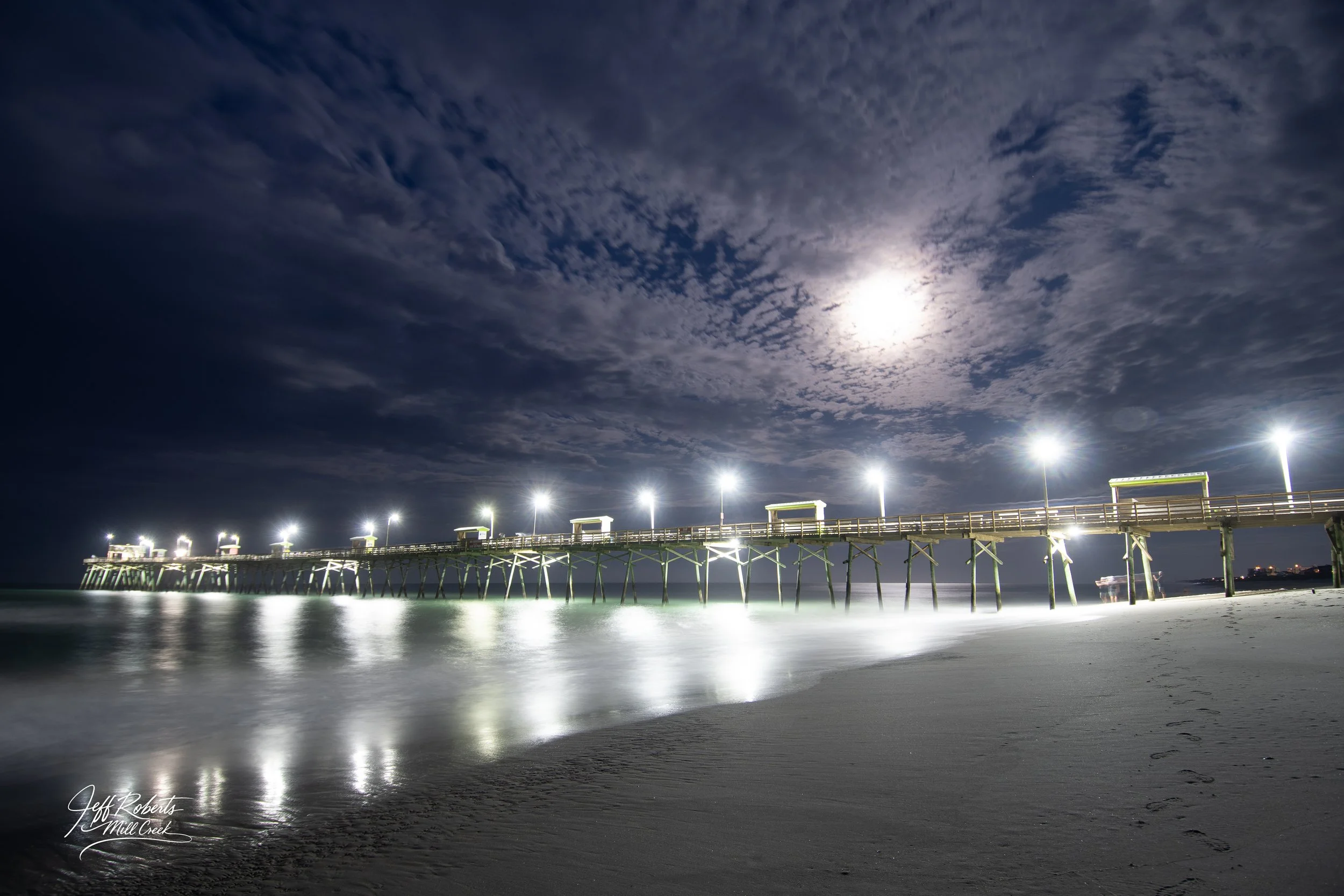 Nighttime view of a wooden pier extending over the ocean, illuminated by bright streetlights, with footprints in the sand and a cloudy sky with the moon shining through.