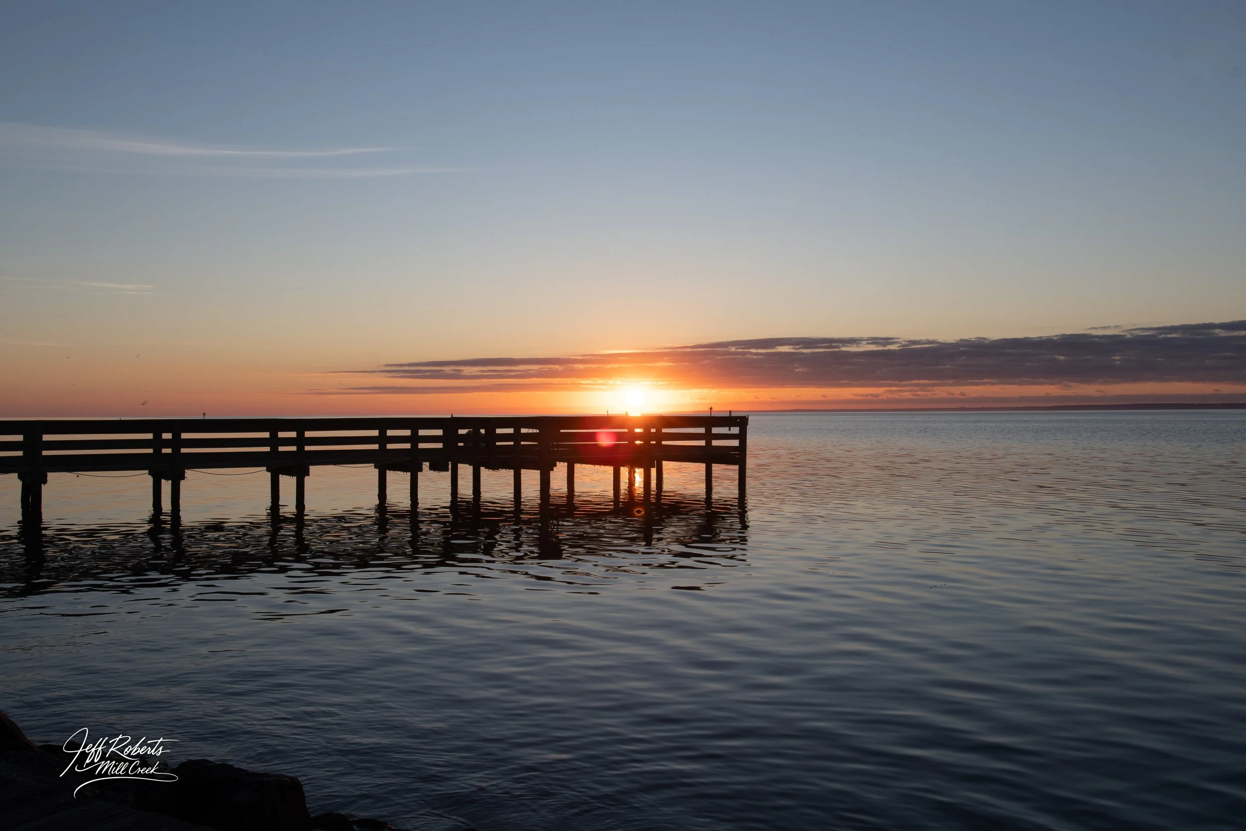 Sunset over a wooden pier extending into calm water with a colorful sky and scattered clouds.