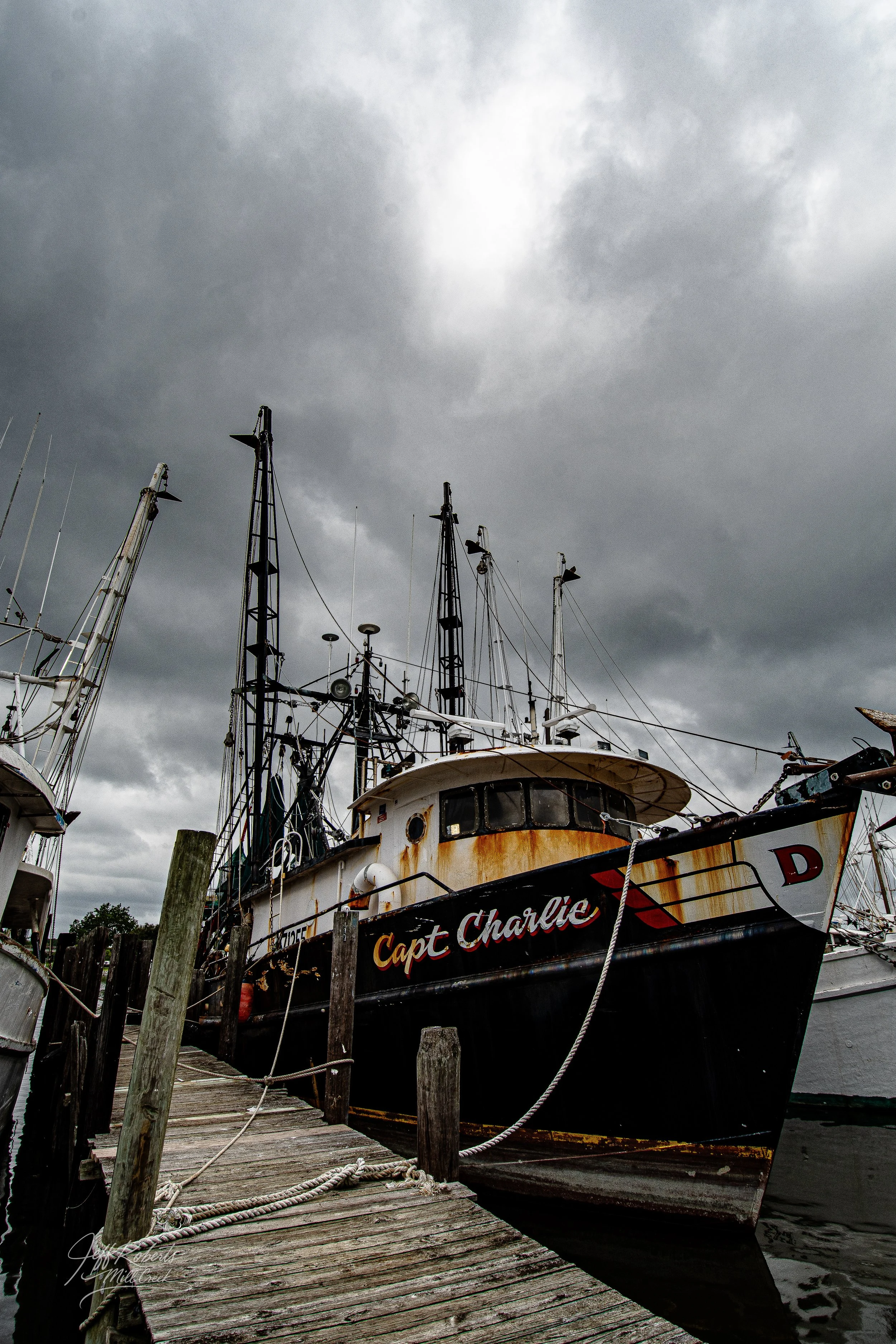 A fishing boat named 'Capt Charlie' docked at a weathered wooden pier on a cloudy day with dark, overcast skies.