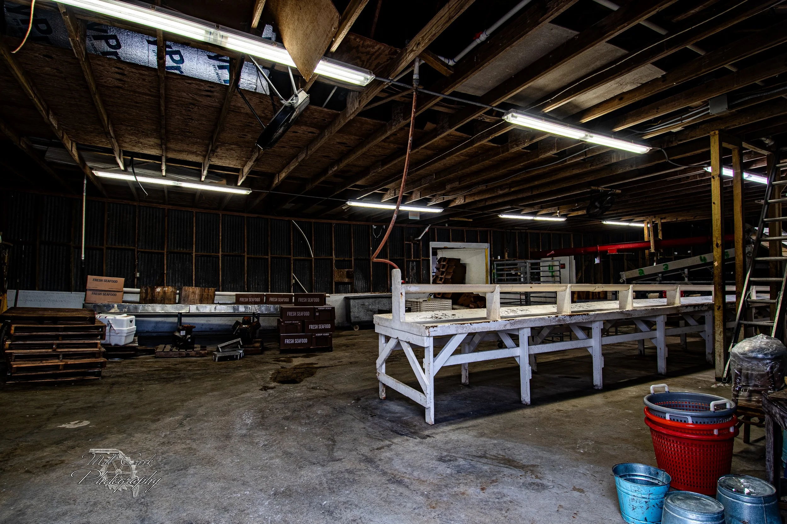 "Empty Packing House"
Inside a weathered seafood packing house, the once-busy sorting tables now stand empty beneath fluorescent lights. Wooden beams, scattered pallets, ice bins, and seafood crates hint at long days of labor gone quiet, preserving t