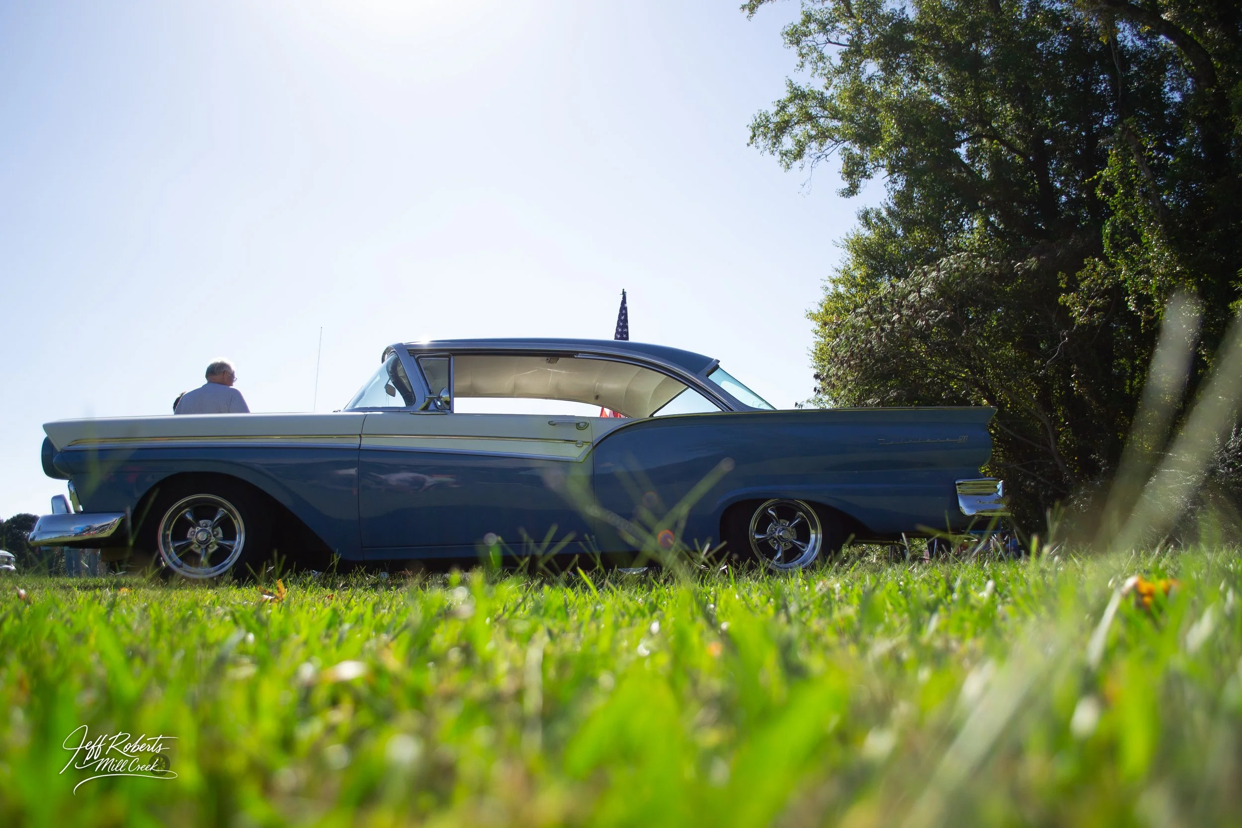 A vintage blue and white car parked on grass with a person standing beside it and trees in the background under a bright blue sky.