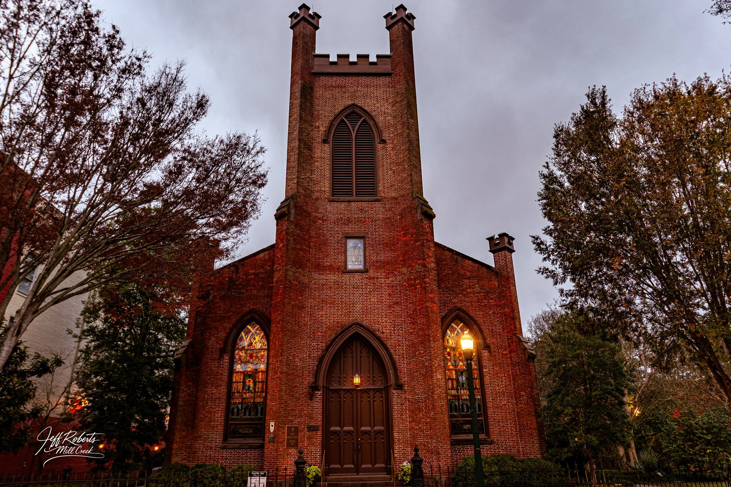 A red brick church with tall, narrow stained glass windows, a large arched wooden door, and a tower with battlements on top, surrounded by trees during overcast weather.