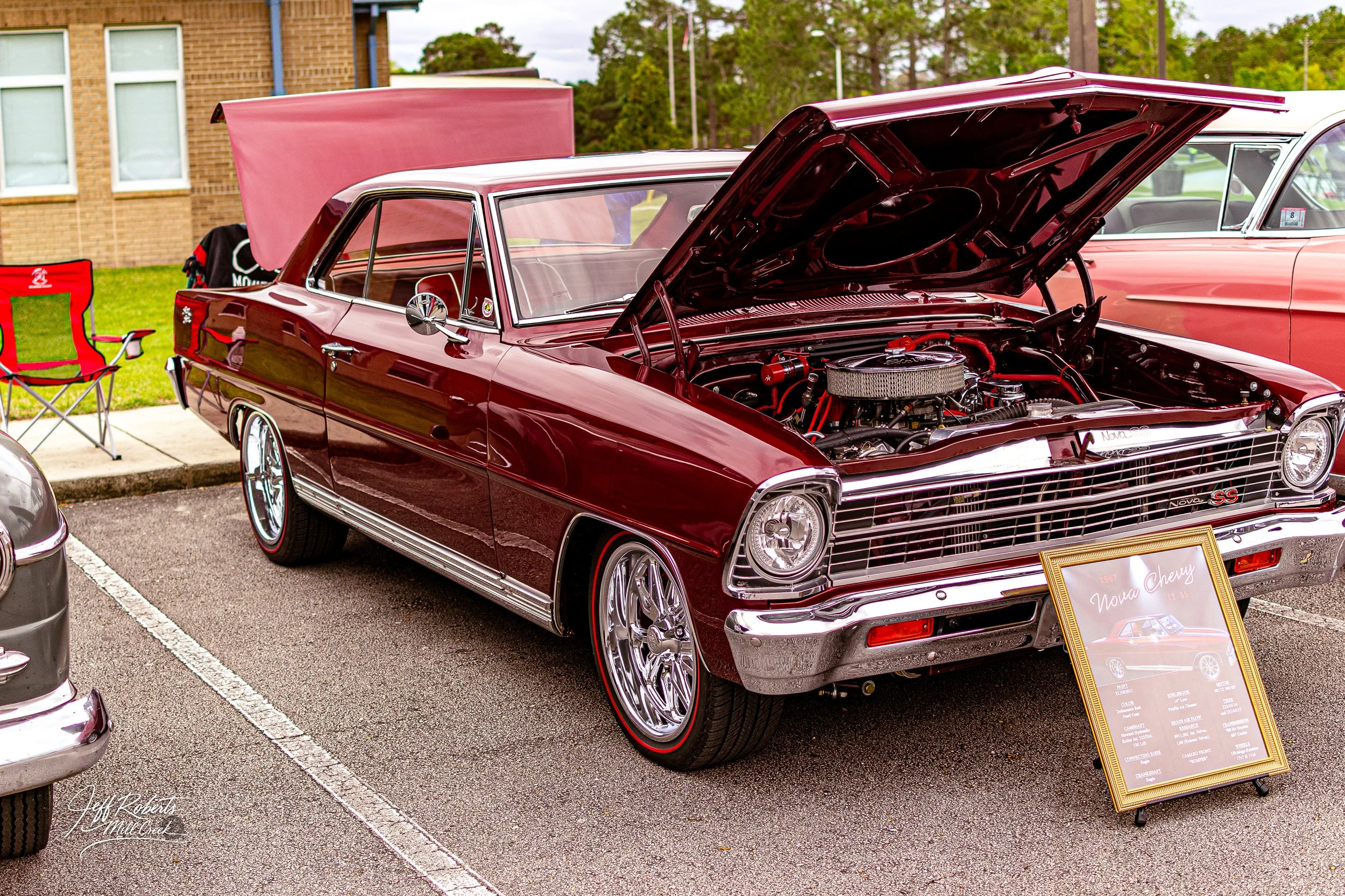 Maroon vintage Chevrolet Nova SS on display at a car show with the hood open, showing its engine. Next to it is a framed display board with details about the car.