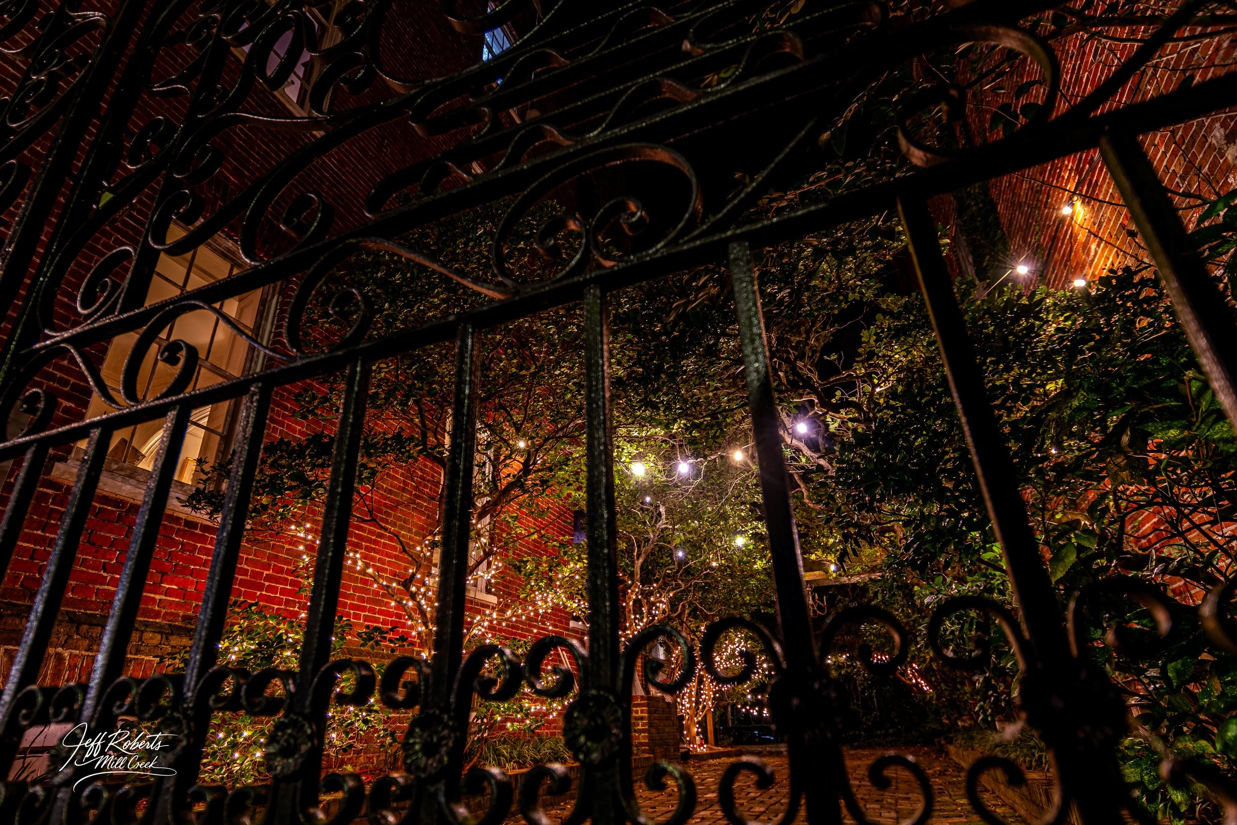 Nighttime view of a brick building with flower boxes and trees decorated with string lights, seen through a decorative black wrought iron fence.
