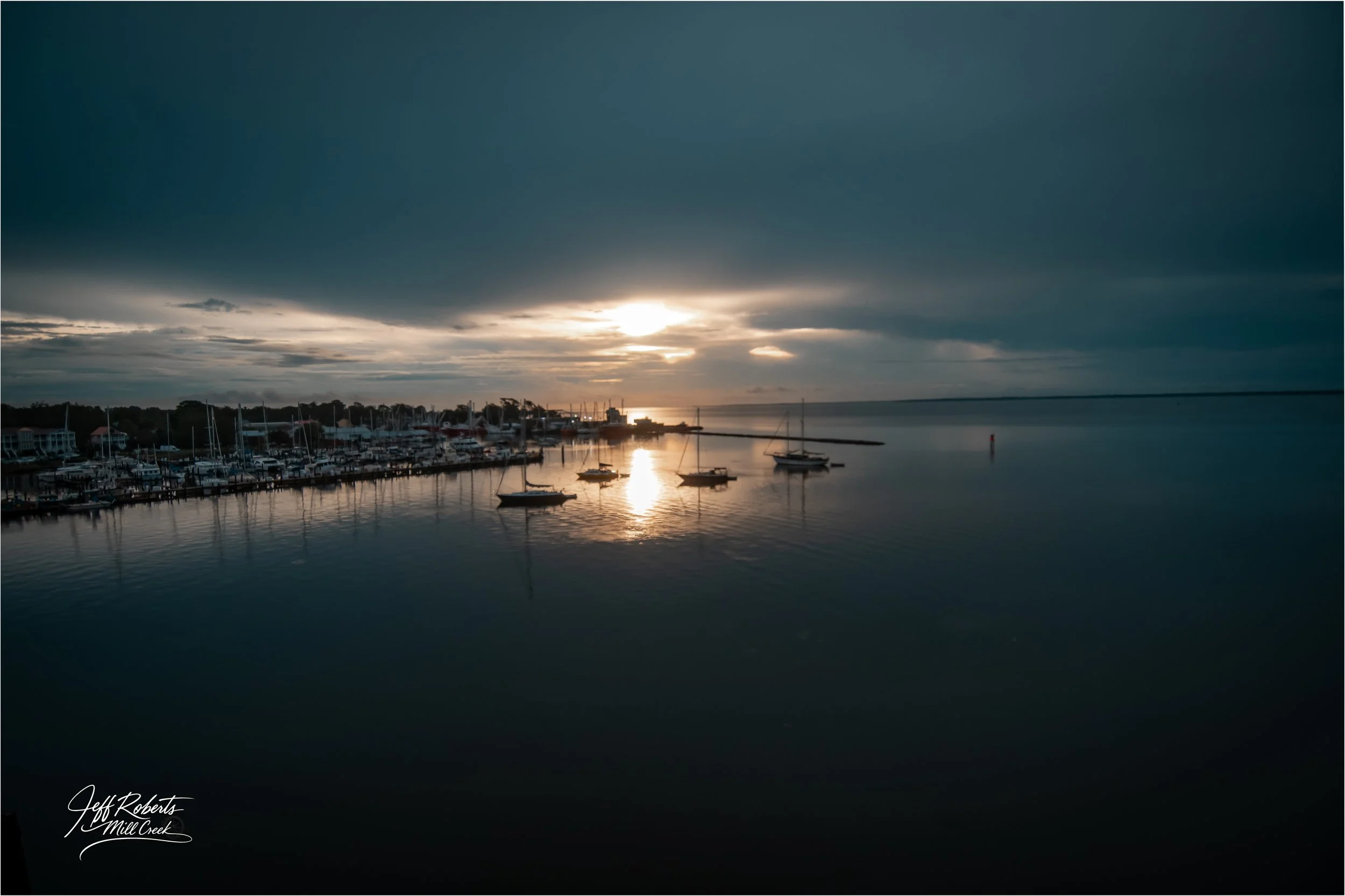 Sunset over a calm marina with several sailboats docked, dark clouds overhead, and a reflection of the sun on the water.