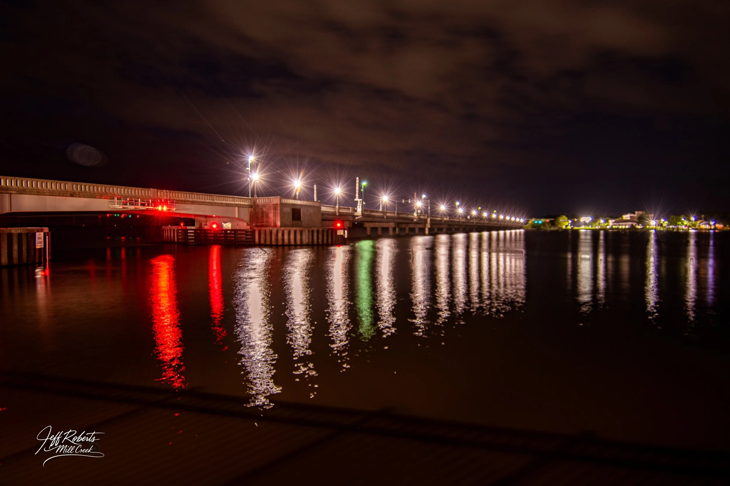 Nighttime view of a bridge over Mill Creek with streetlights reflecting in the water, and a dark cloudy sky.